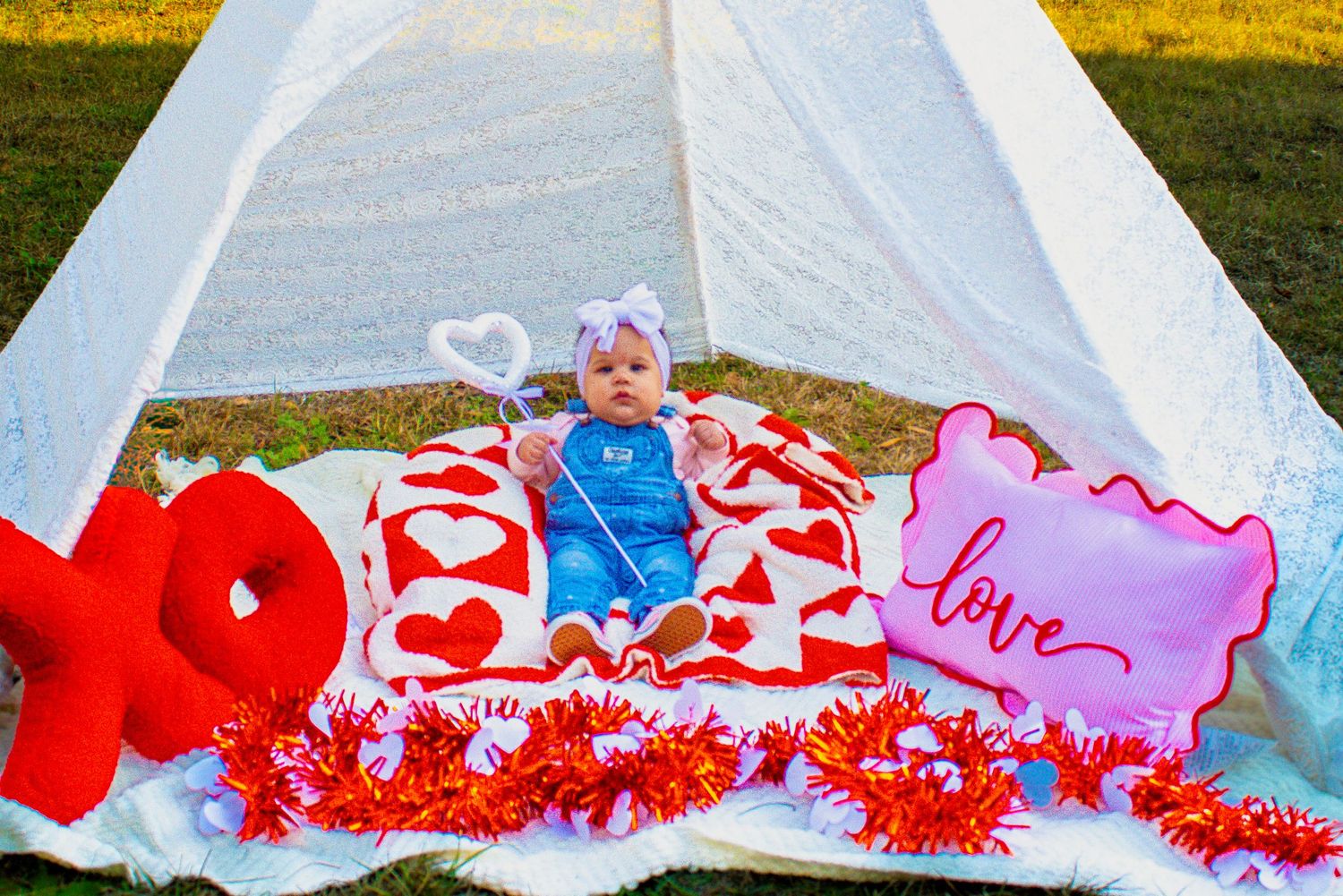 Baby in blue outfit with white bow sits on heart blanket near white teepee with XO and love signs.