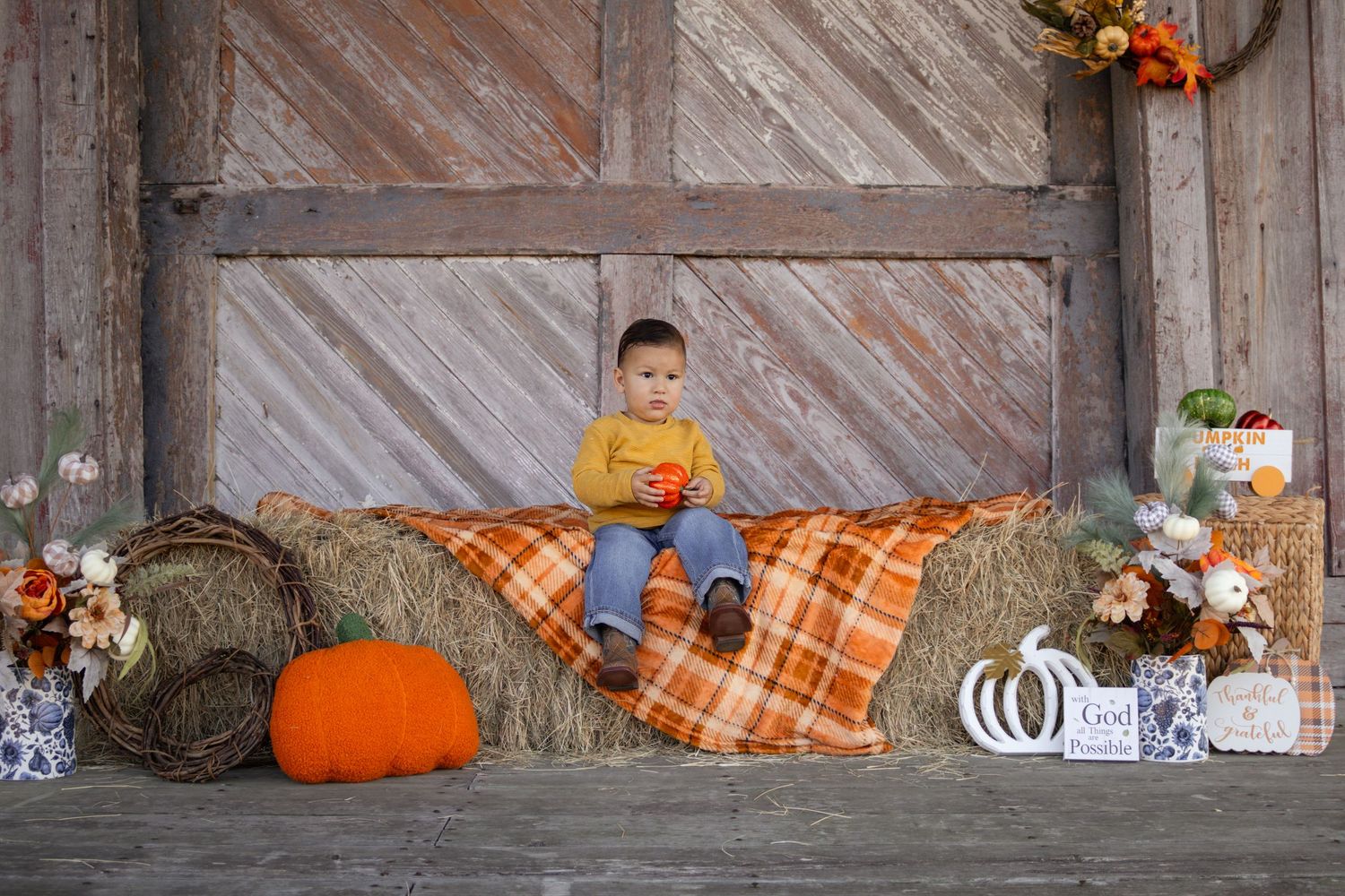 Young boy in yellow sweater sitting on plaid blanket on hay bale with fall pumpkin decor.