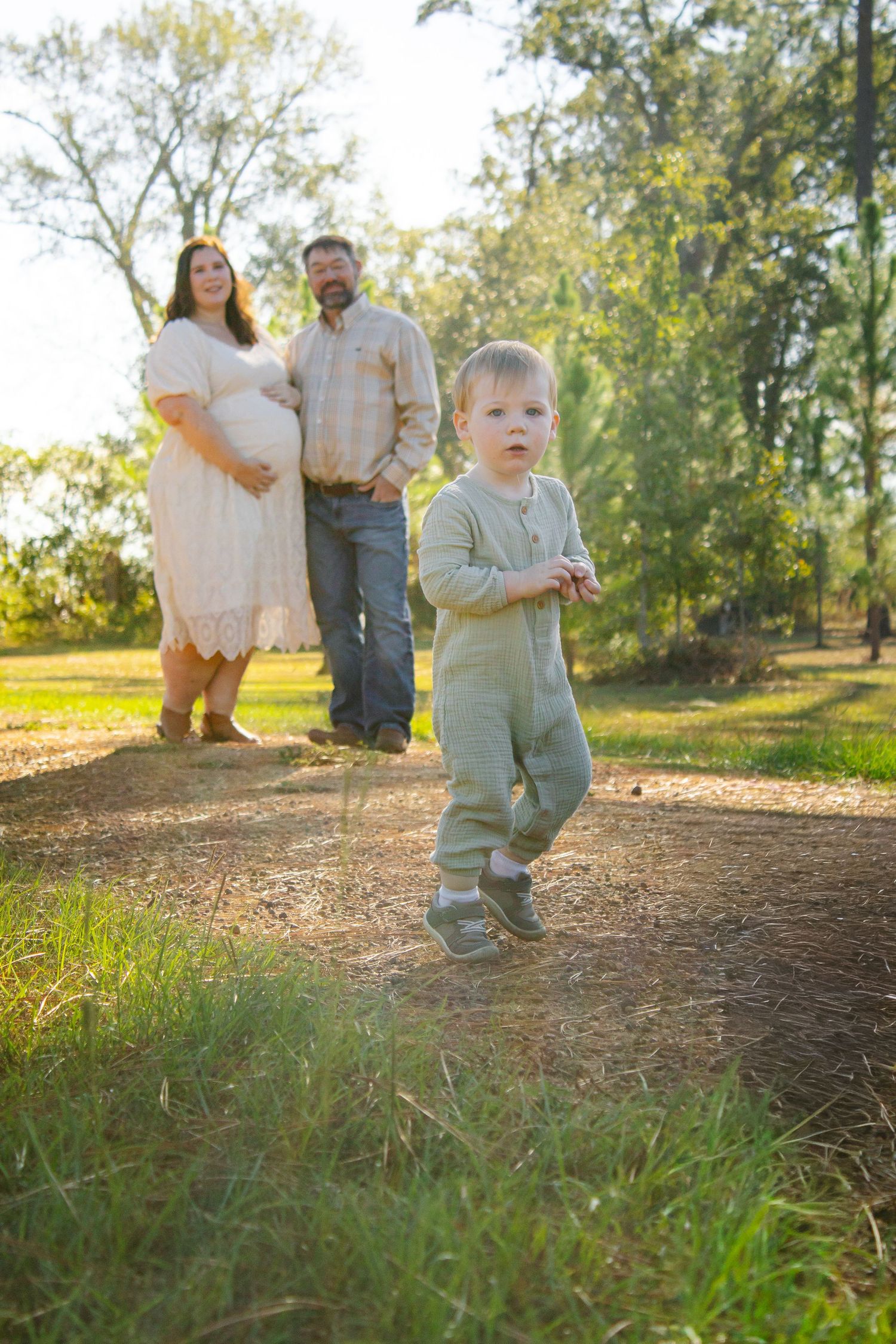 Family maternity photo with toddler running on path as parents stand in background.
