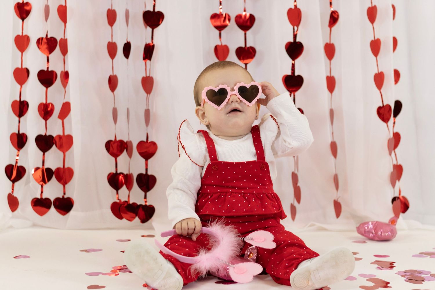 Adorable baby in red outfit wears heart-shaped sunglasses surrounded by hanging heart garlands and confetti.
