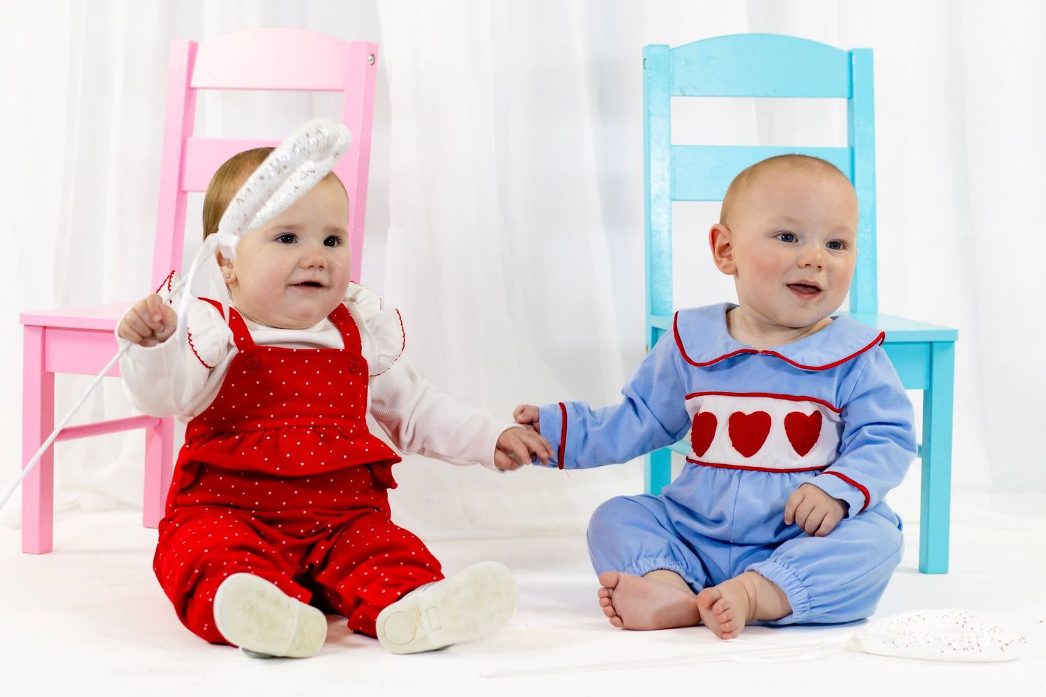 Two babies in Valentine's Day outfits hold hands while sitting in front of pink and blue chairs.