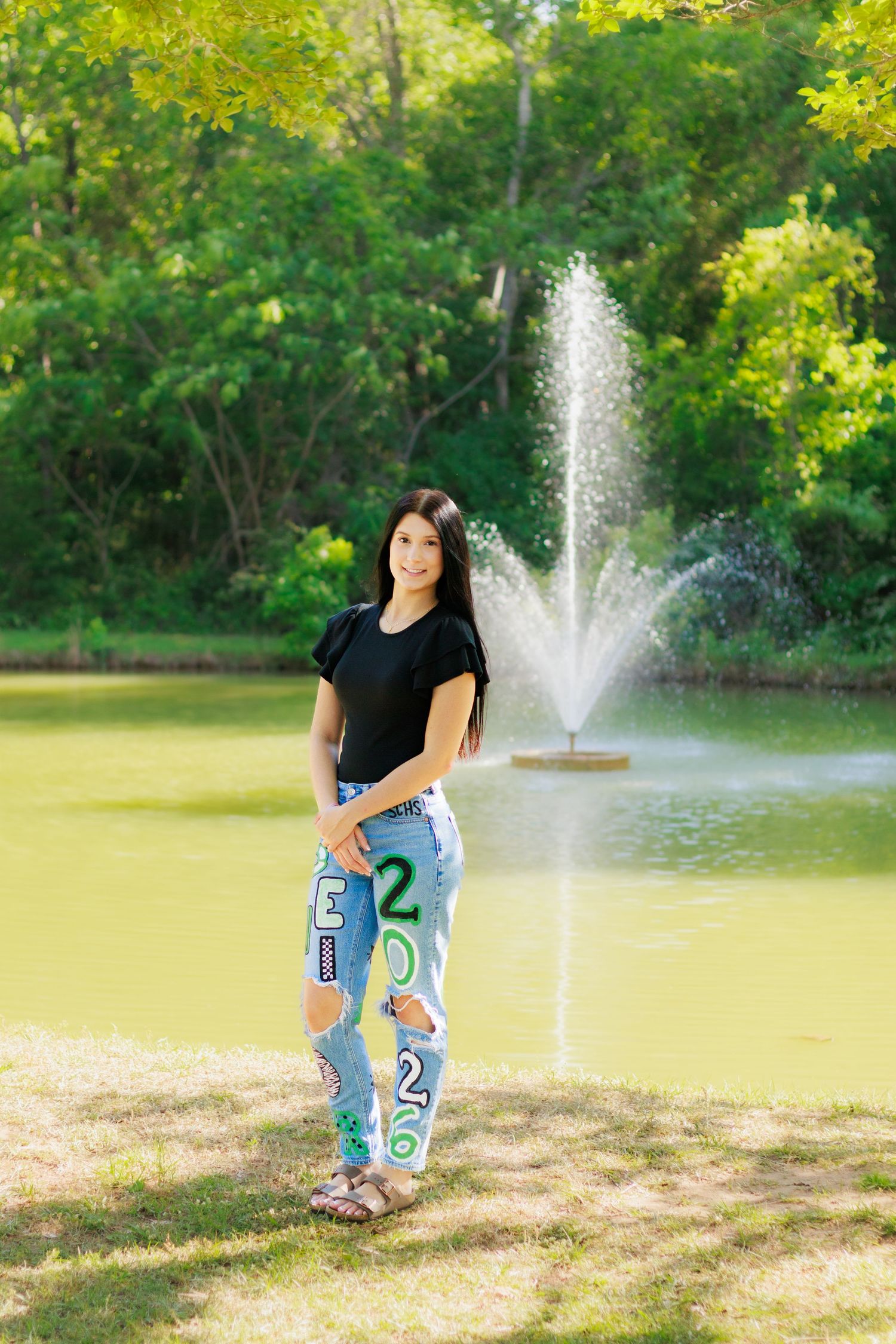 Young woman in decorated graduation jeans stands smiling by a park fountain with lush green trees.