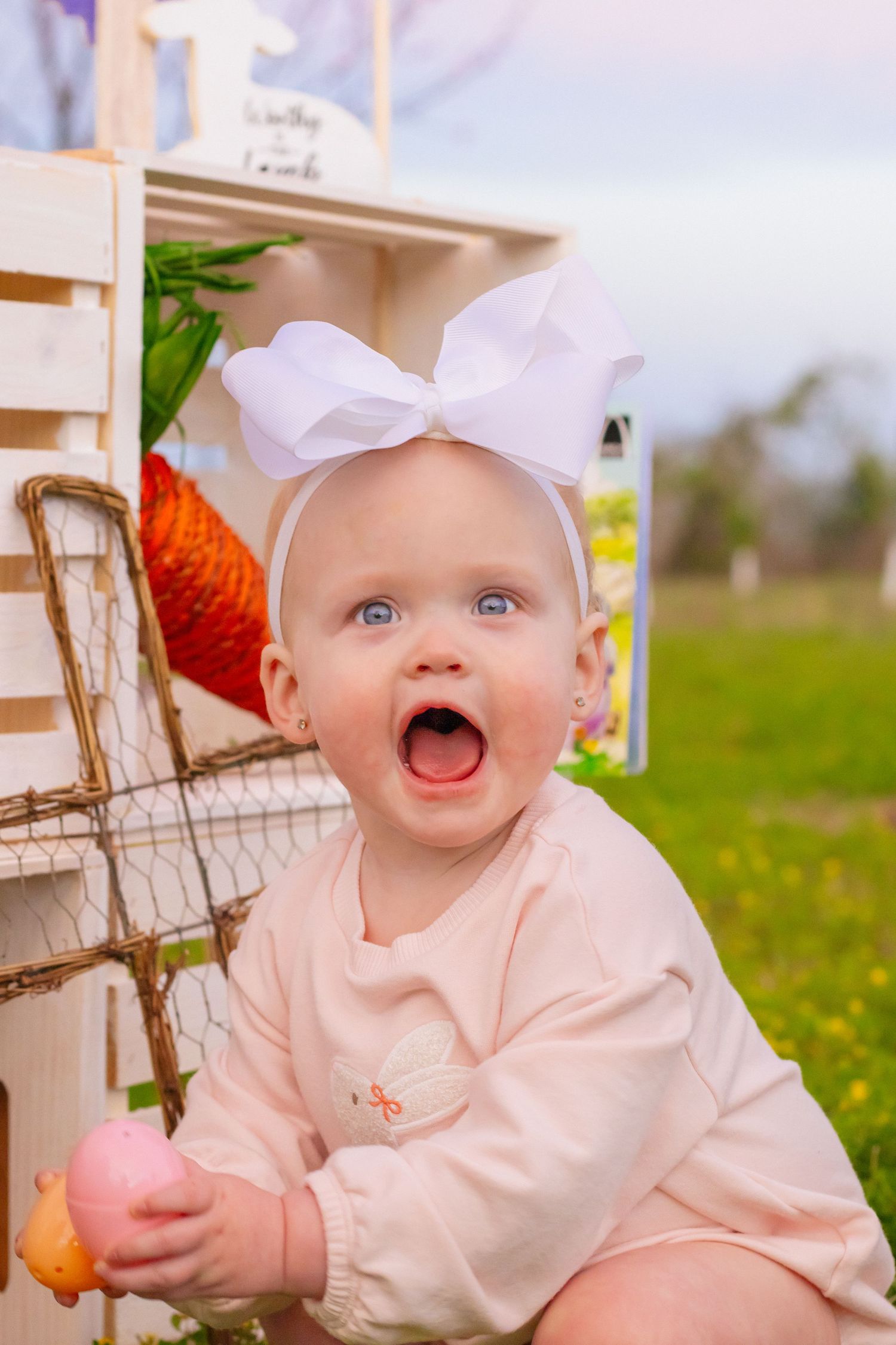 Surprised blue-eyed baby with white bow headband holds Easter egg outdoors.