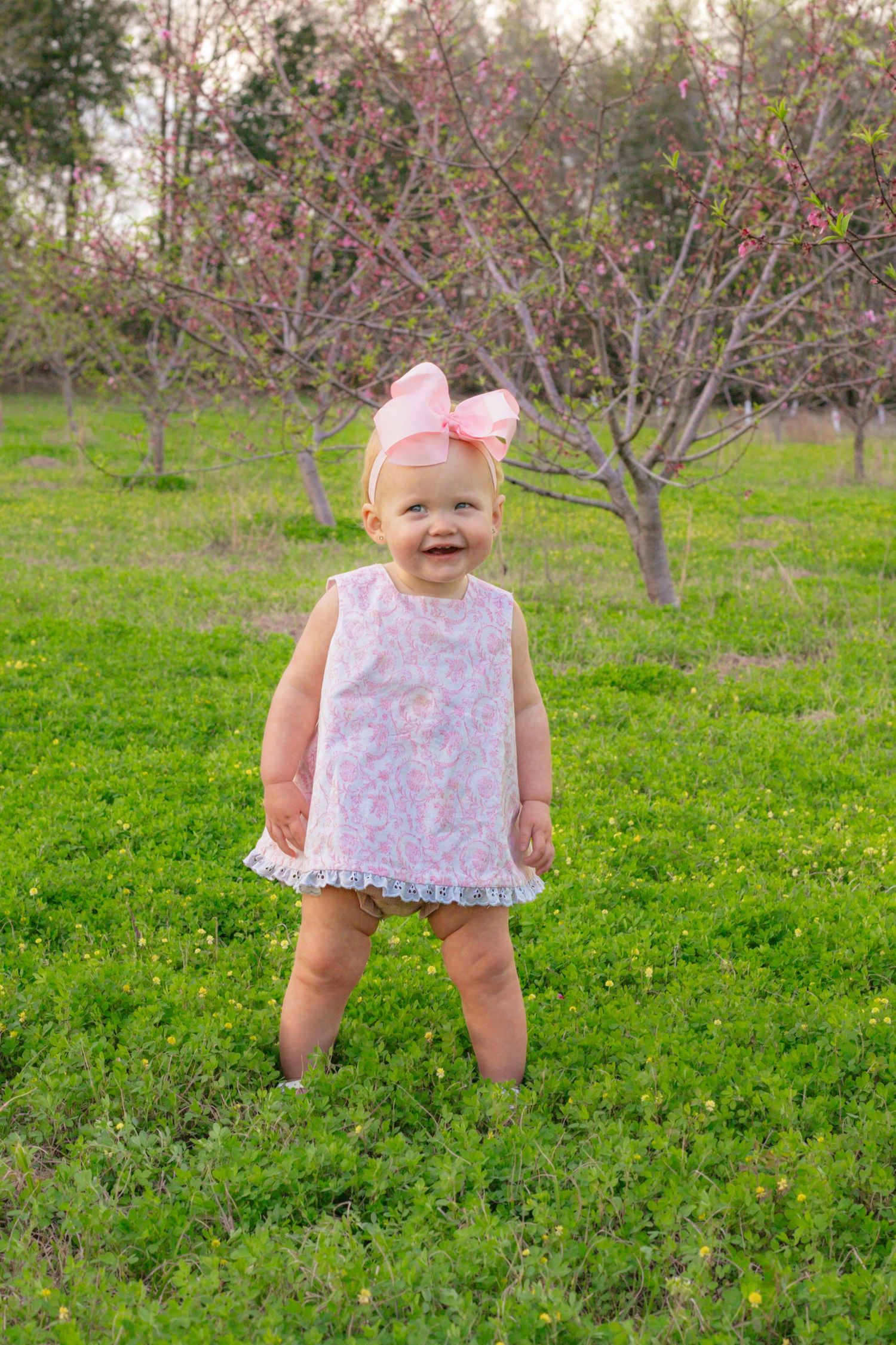 Happy toddler girl wearing pink floral dress and large pink bow headband smiling outdoors in spring orchard.