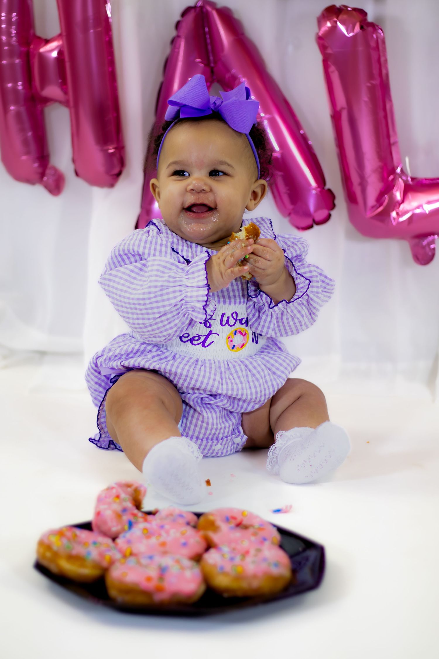 Smiling baby girl in purple gingham romper enjoys donut at half birthday photo shoot.