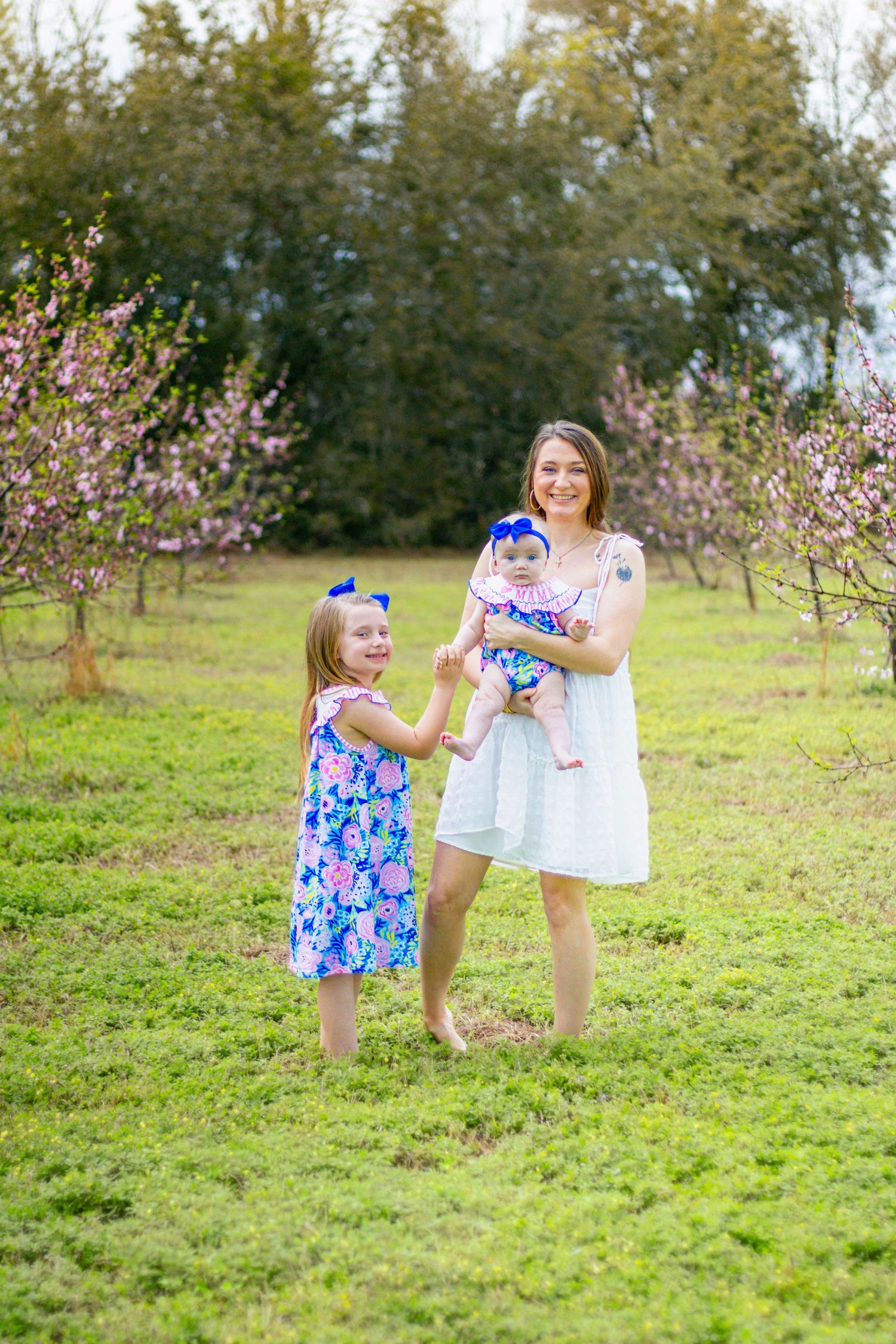 Mother in white dress holding baby while toddler in blue floral dress stands in blooming orchard.