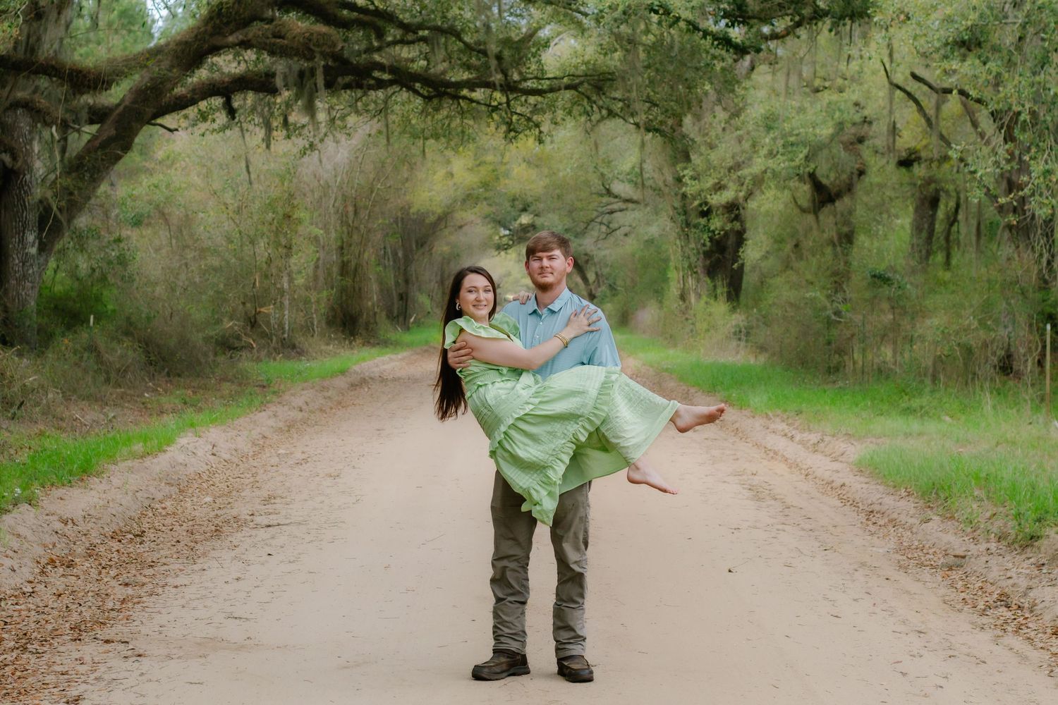 Man carrying woman in green dress in his arms on a beautiful tree-lined dirt road.