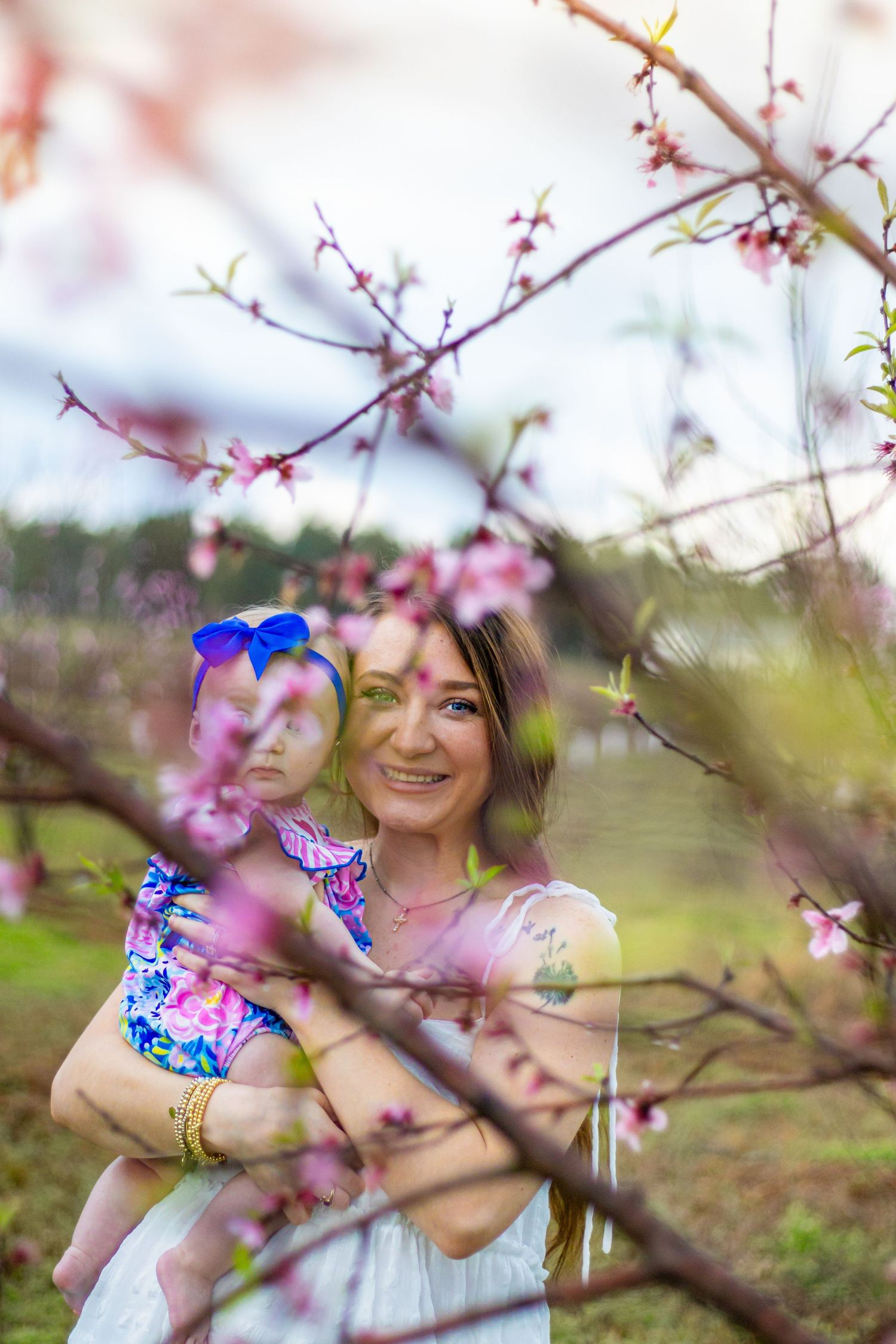 Mother cradles infant daughter with blue headband among blooming pink cherry blossom trees outdoors.