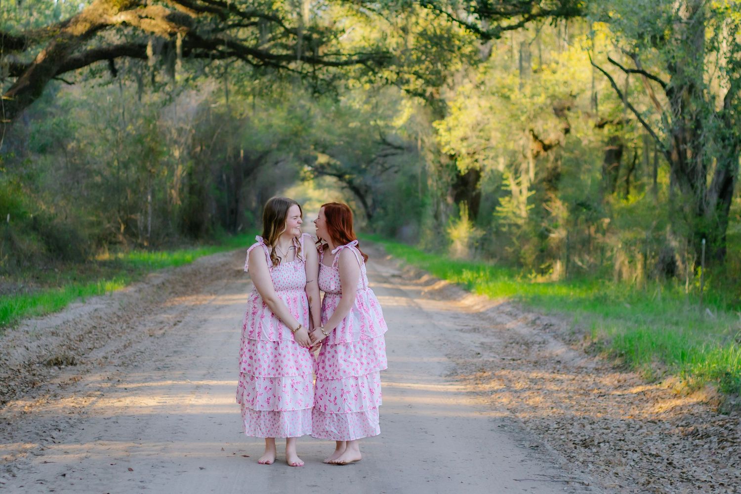 Two girls wearing matching pink dresses stand facing each other on a sunlit dirt path.