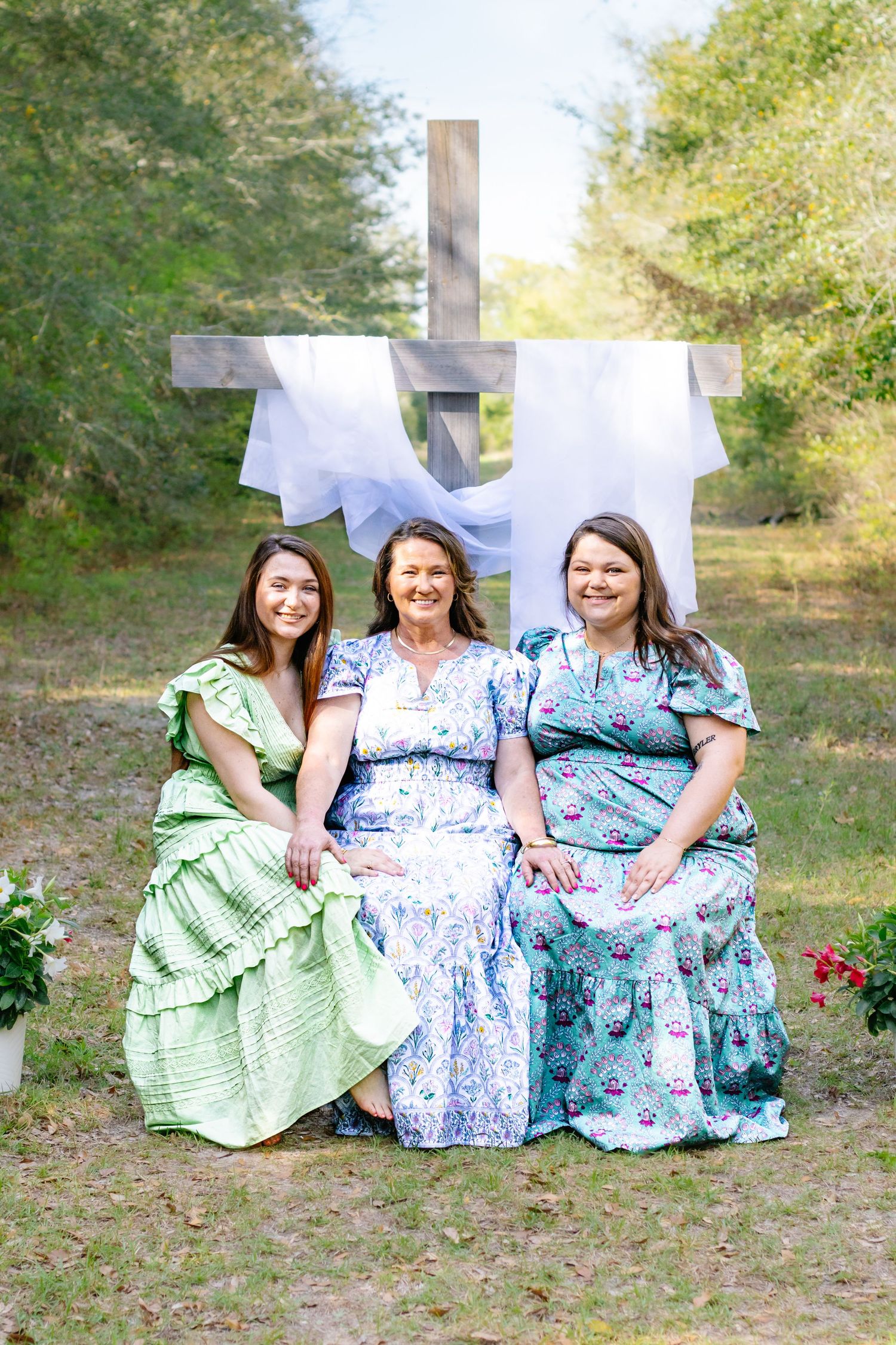 Three women in floral dresses seated together outdoors with wooden cross and white drape backdrop.