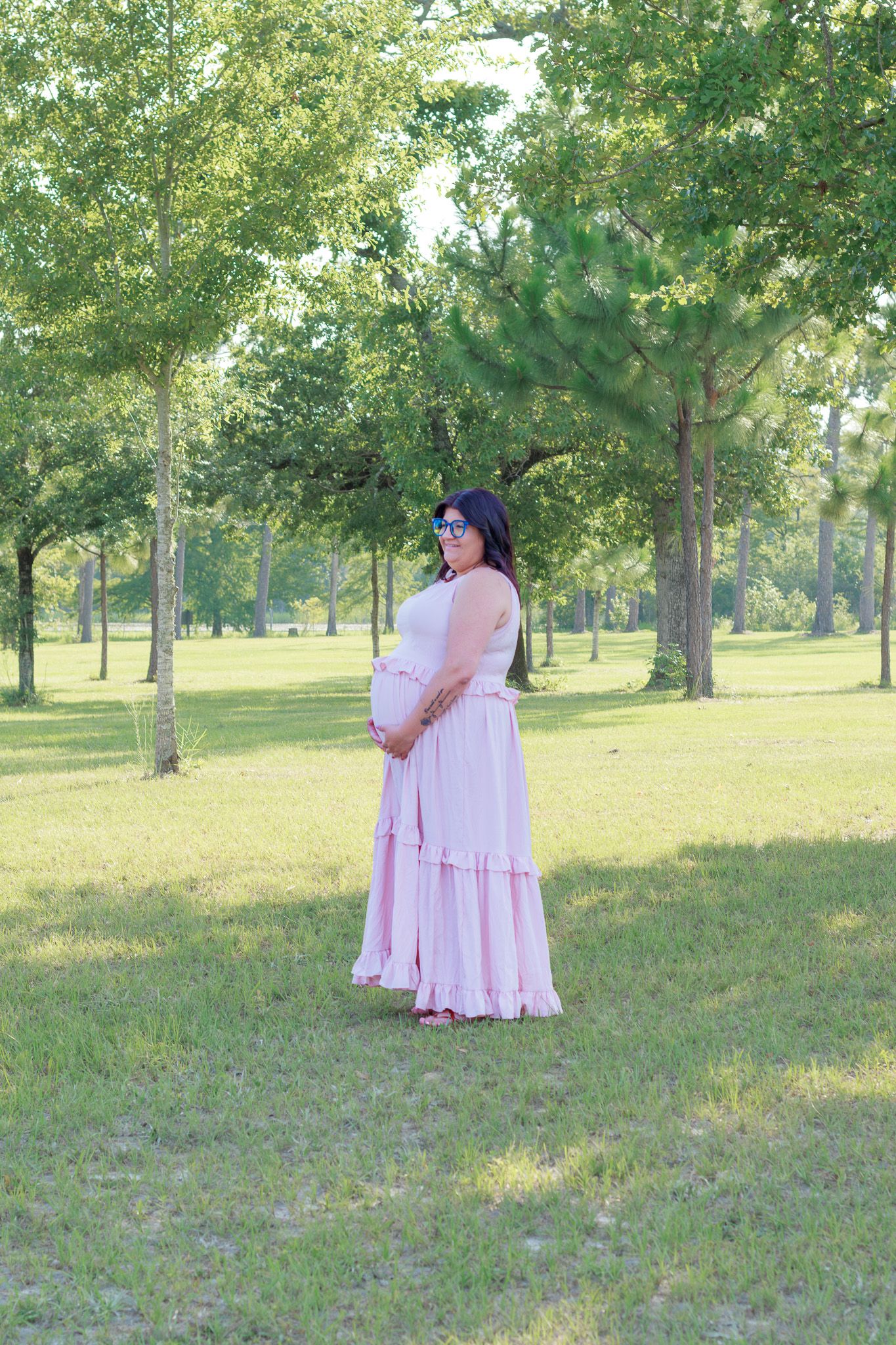 Pregnant woman in pink maxi dress posing outdoors in a lush green park, cradling her baby bump.