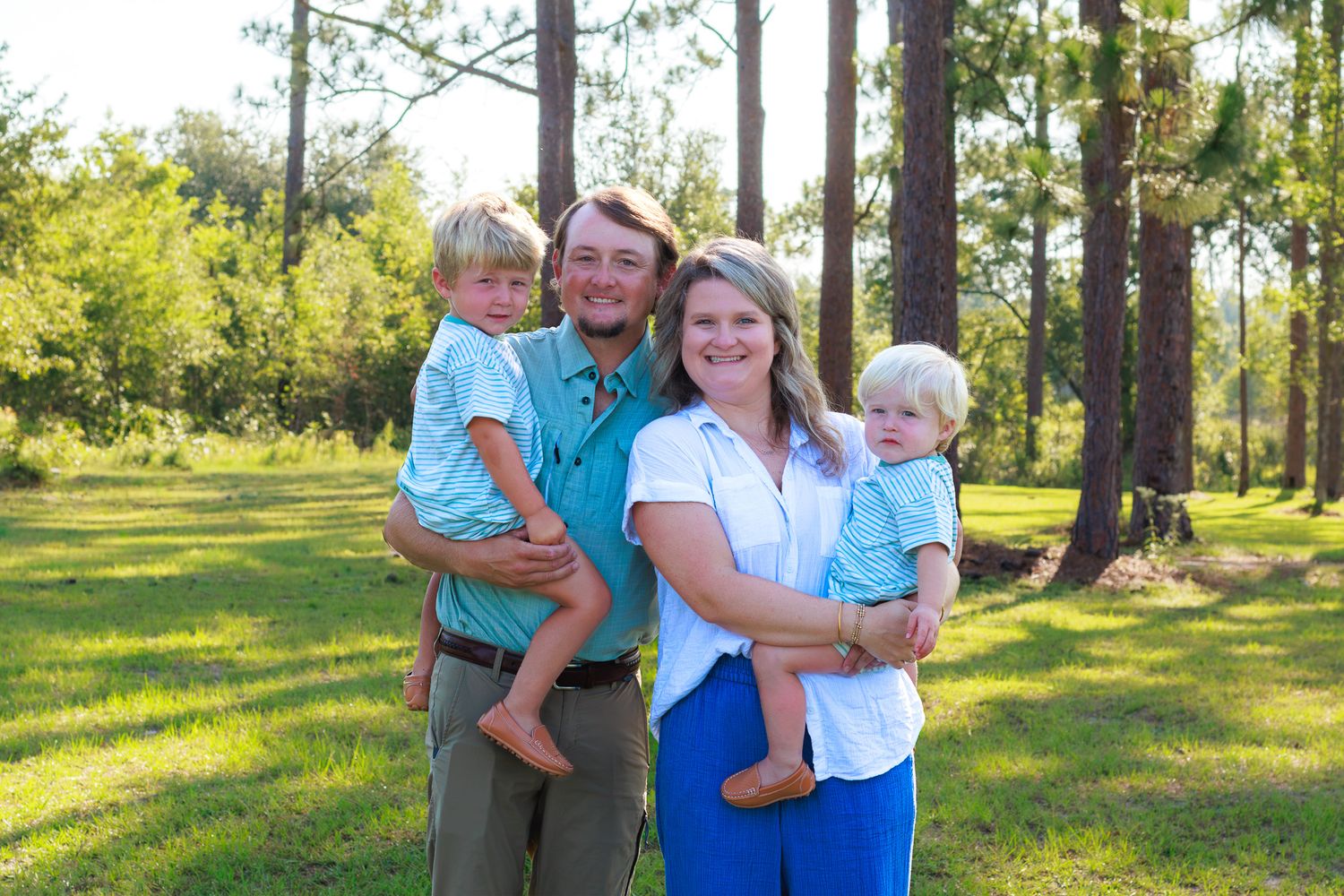 A happy family of four poses outdoors among tall pine trees on a sunny day in a green grassy field.