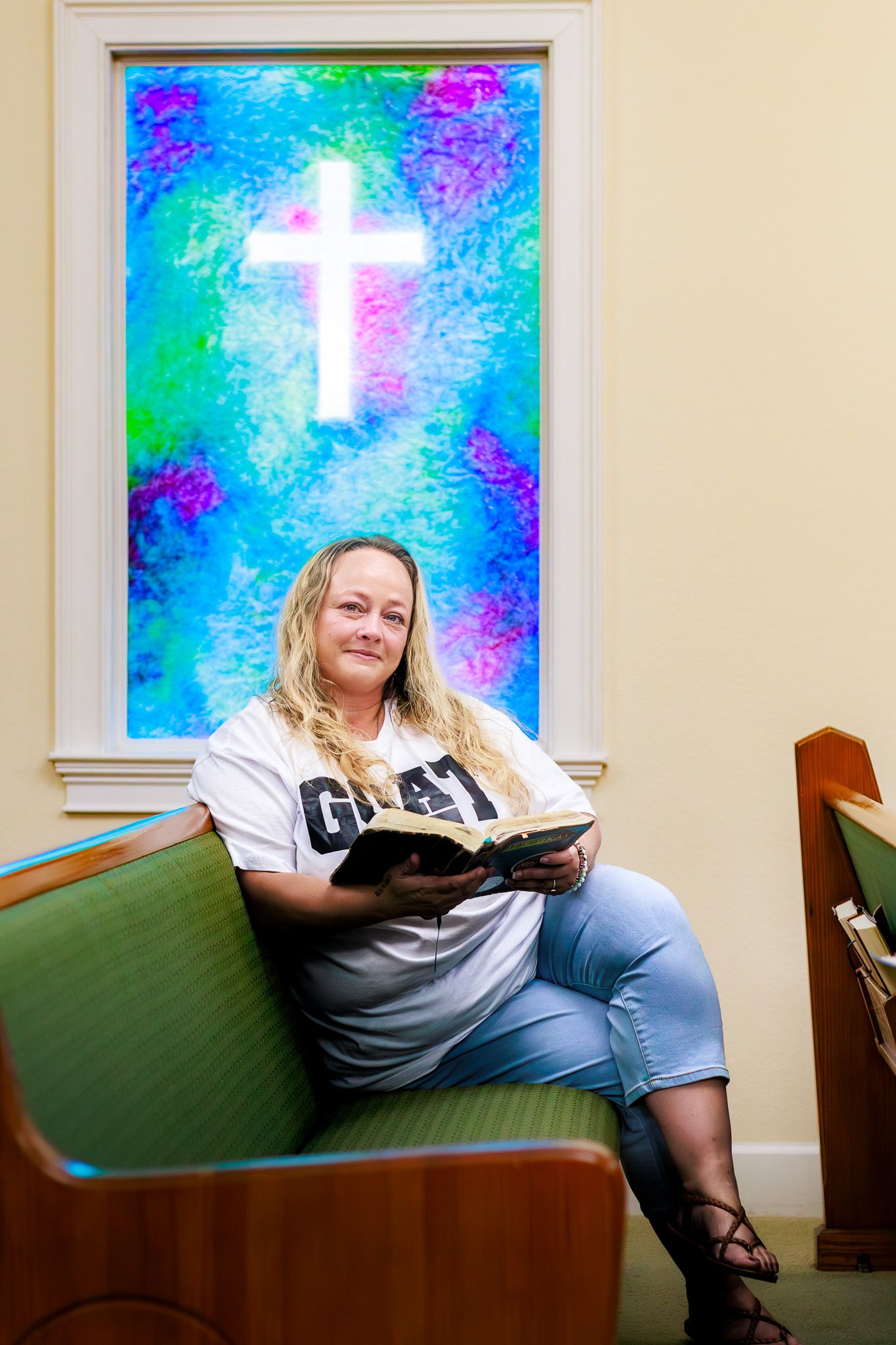 Woman reading Bible seated in church pew before colorful stained glass window with cross.