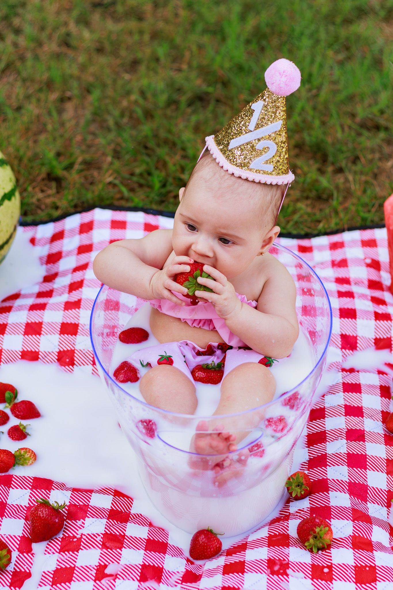 Baby in milk bath with strawberries, wearing gold birthday crown, sitting in glass bowl on red checkered blanket outdoors.