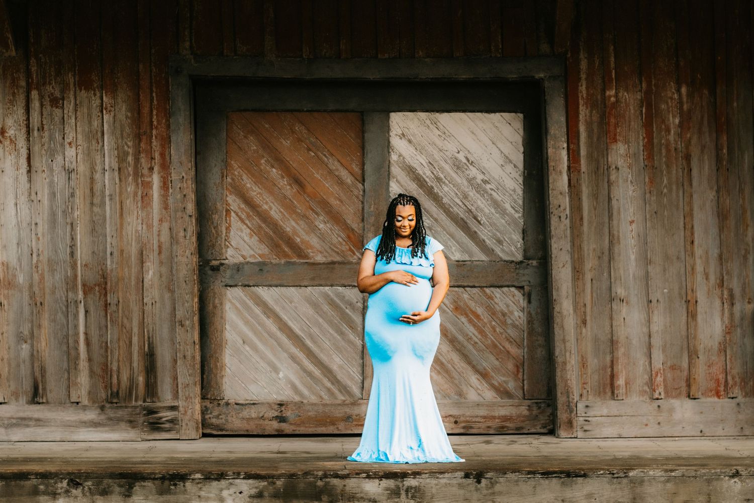 Pregnant woman in a light blue mermaid gown cradling her baby bump, standing in front of a rustic wooden barn.