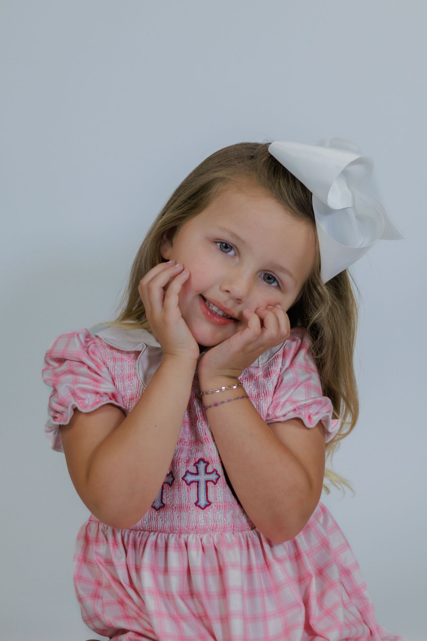 Adorable young girl in pink gingham smocked dress with white bow, posing with hands on cheeks and smiling.