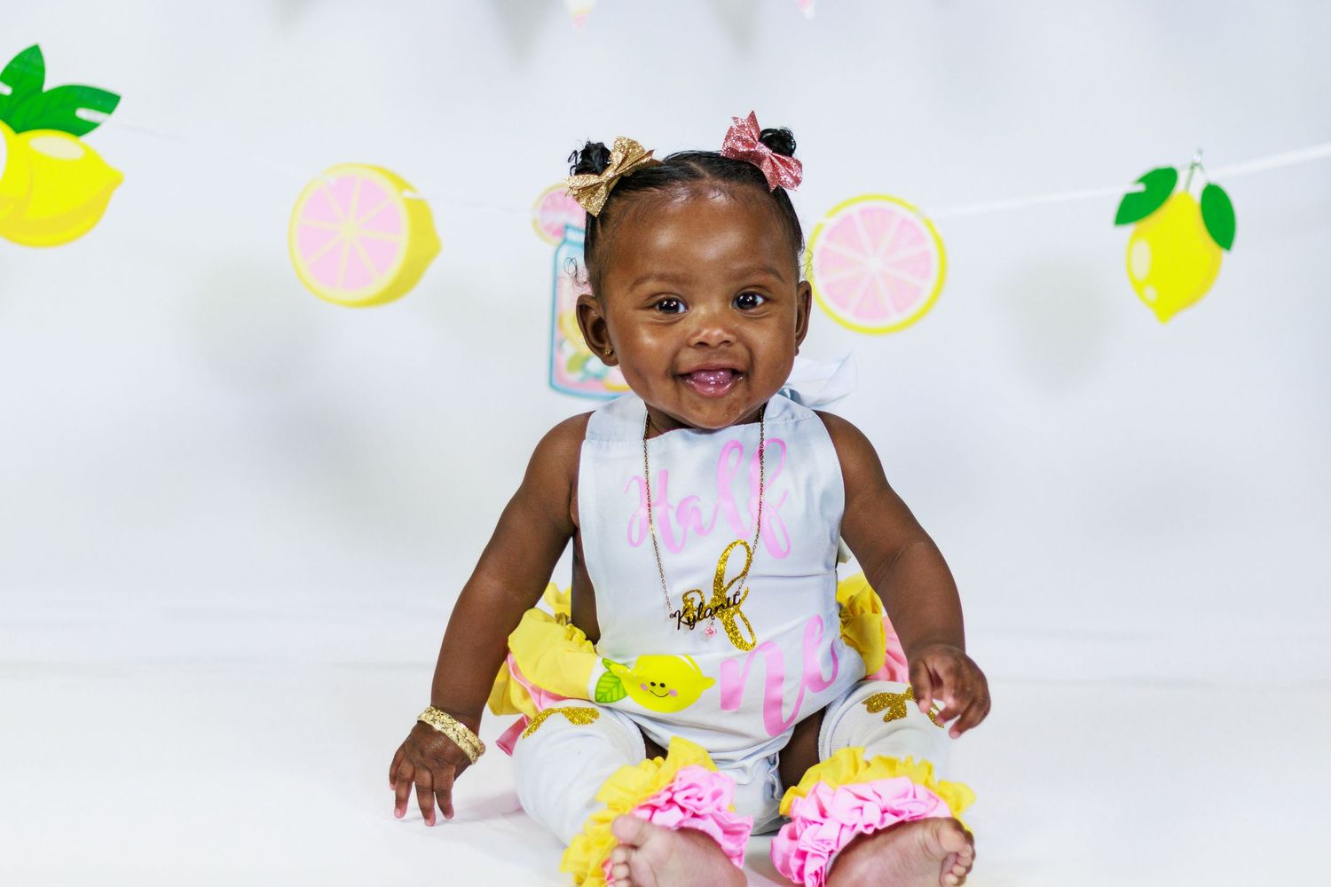 Smiling baby girl in lemon print romper with ruffled skirt sitting by citrus party banner.