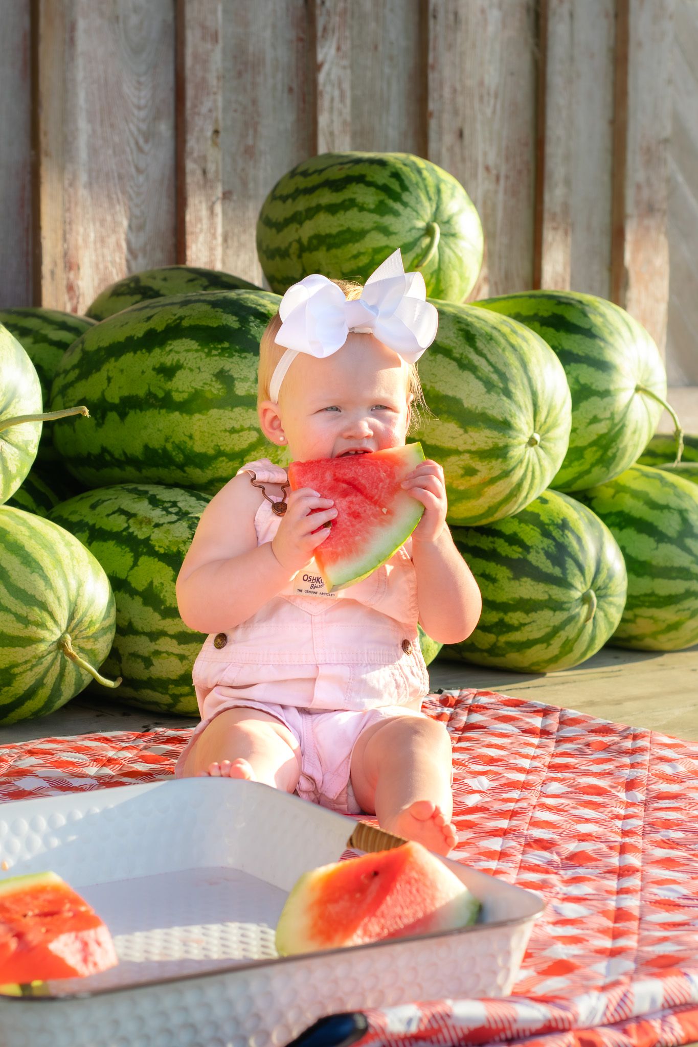 Baby girl in pink outfit eating watermelon slice, sitting in front of stacked watermelons on a red checkered blanket.