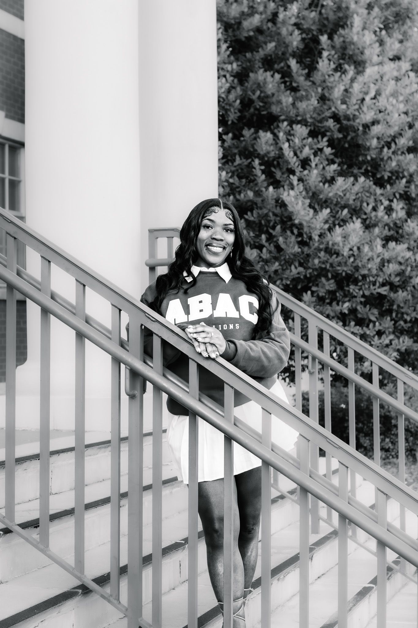 Smiling young woman in ABAC sweatshirt poses on staircase railing in black and white photo.