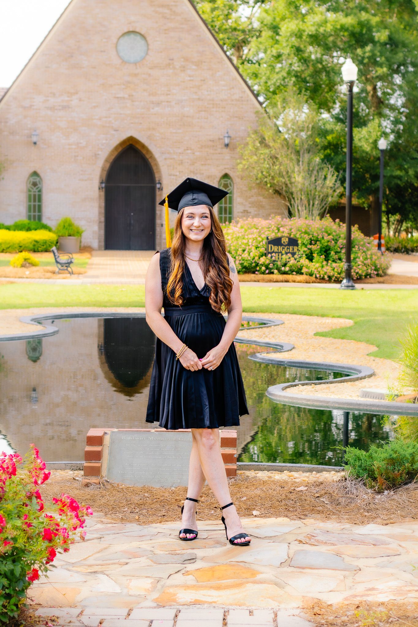 Smiling graduate in black dress and cap stands by reflective pond near chapel building.