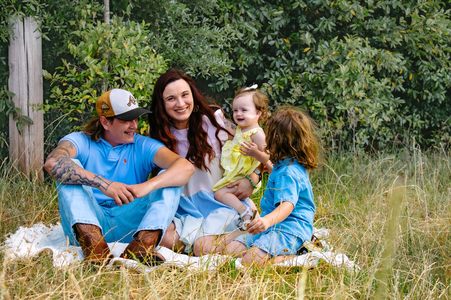 Family of four sitting on a blanket in a grassy field, smiling and enjoying time together outdoors.