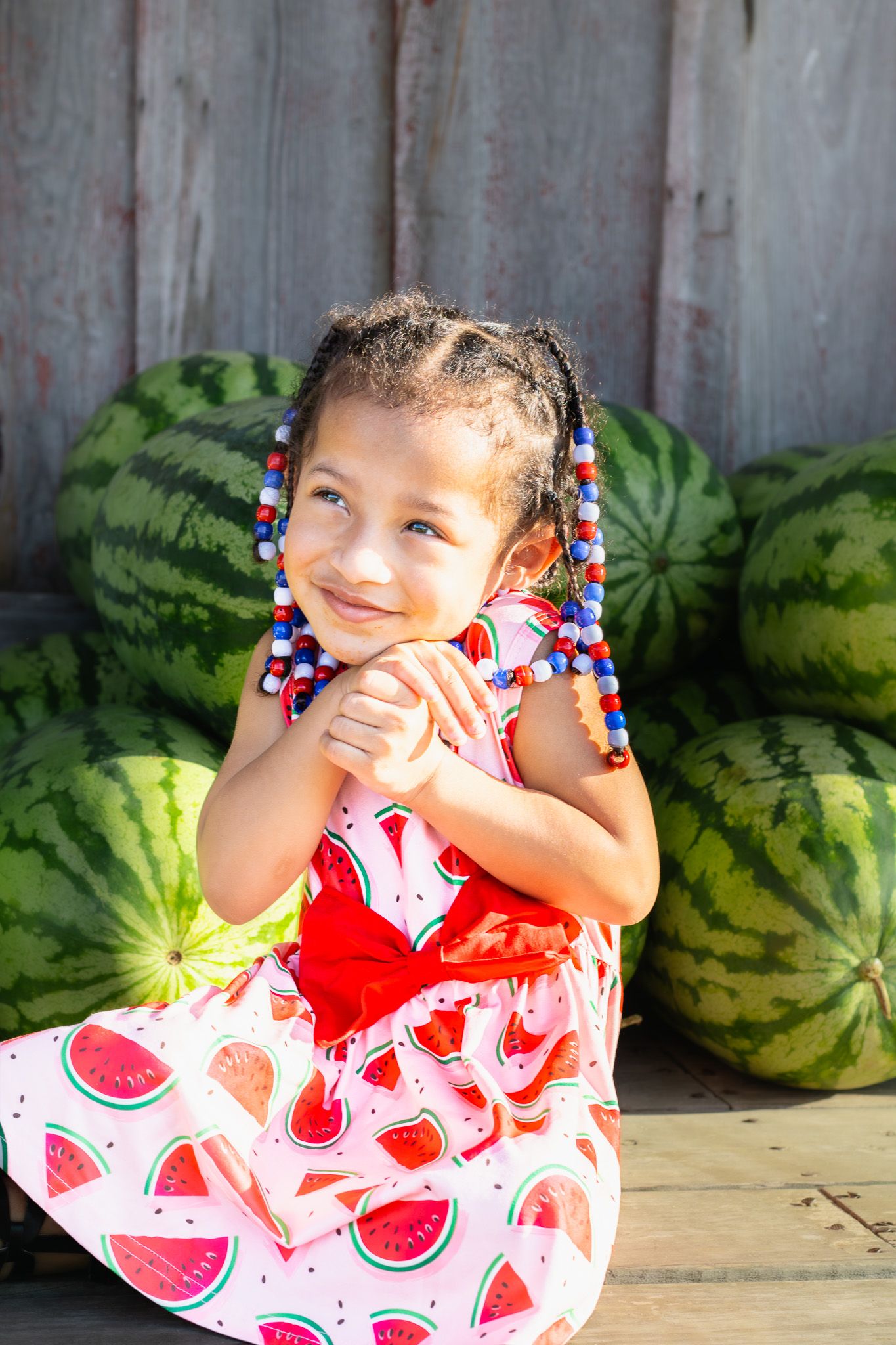 Young girl in watermelon dress with colorful beaded braids smiling beside a pile of watermelons.