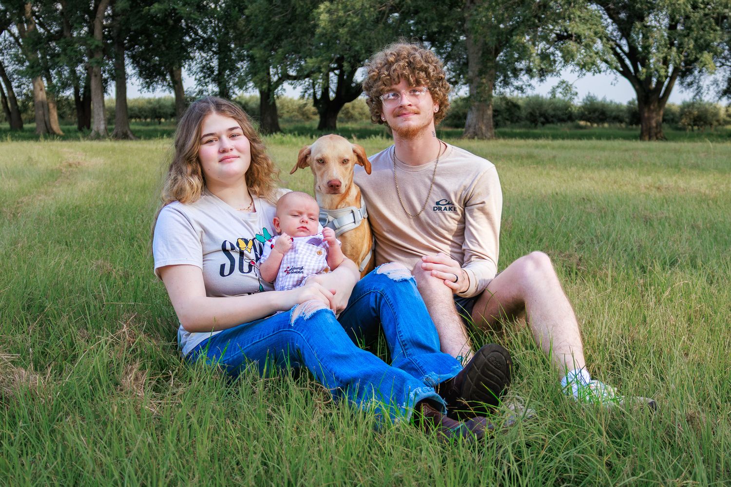Two teens sit in green grass with a baby and brown dog during an outdoor family photo session.