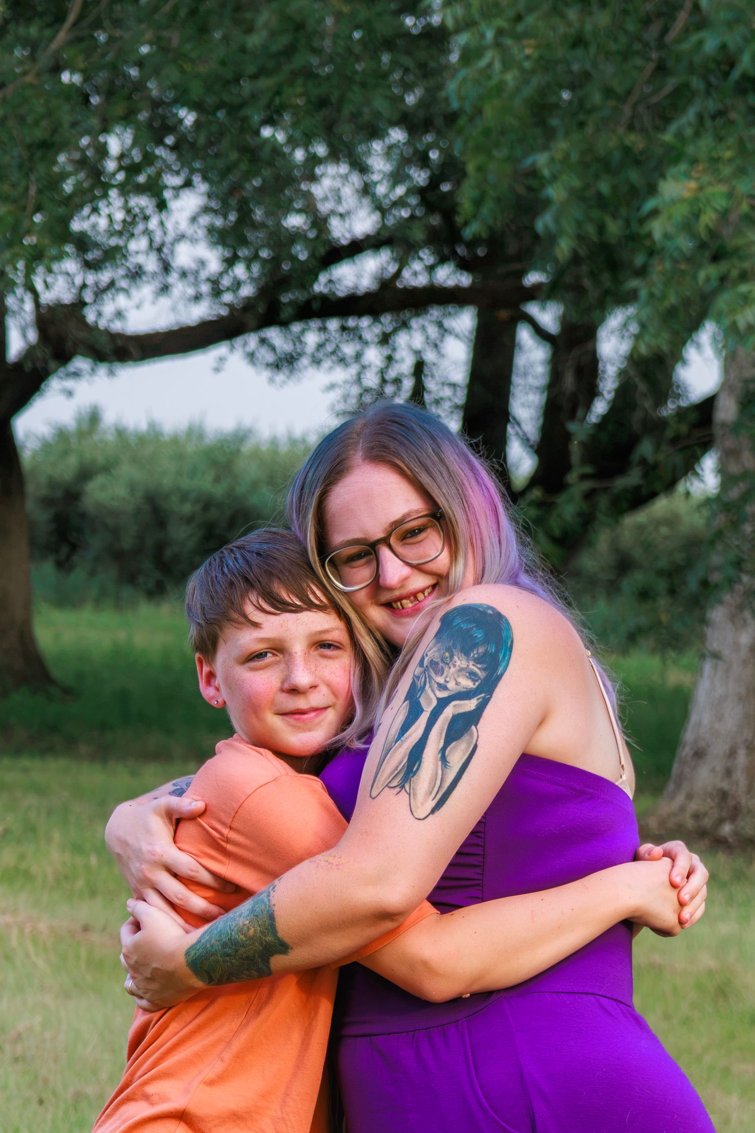 A woman with pink hair and glasses hugs a young boy outdoors, both smiling, with green trees in the background.