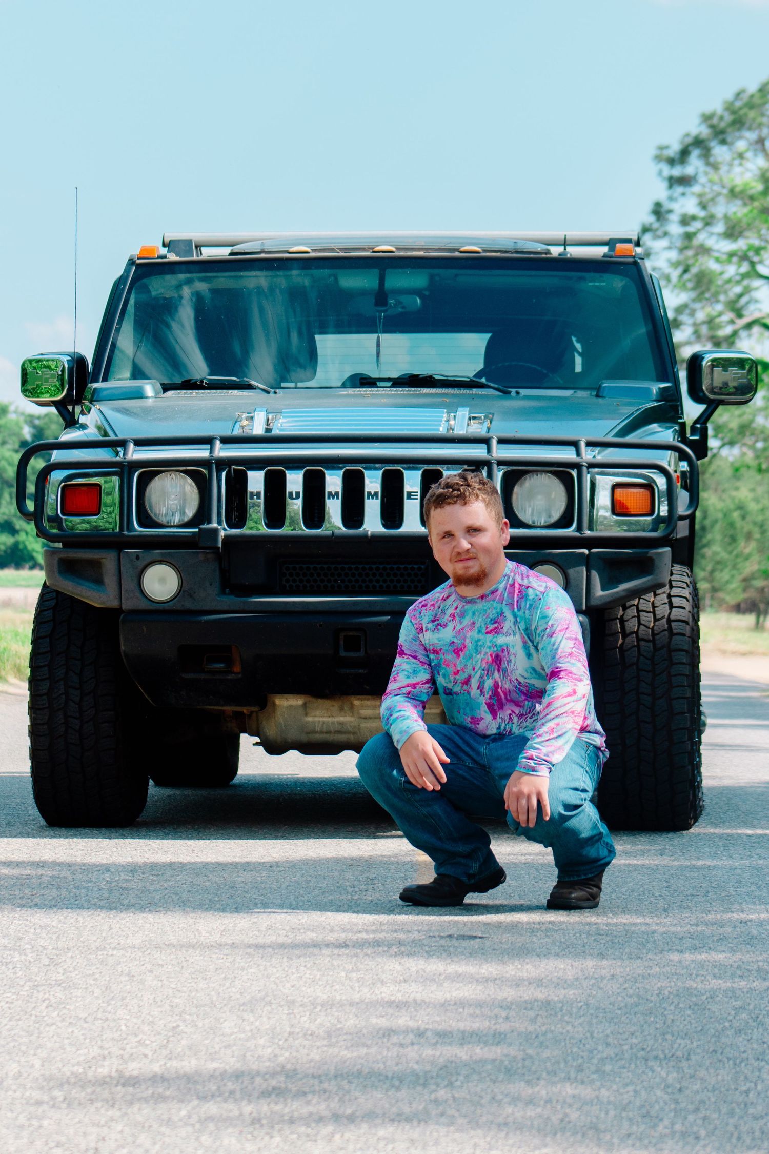 Man in tie-dye shirt crouching in front of a black Hummer H2 on a sunny road.