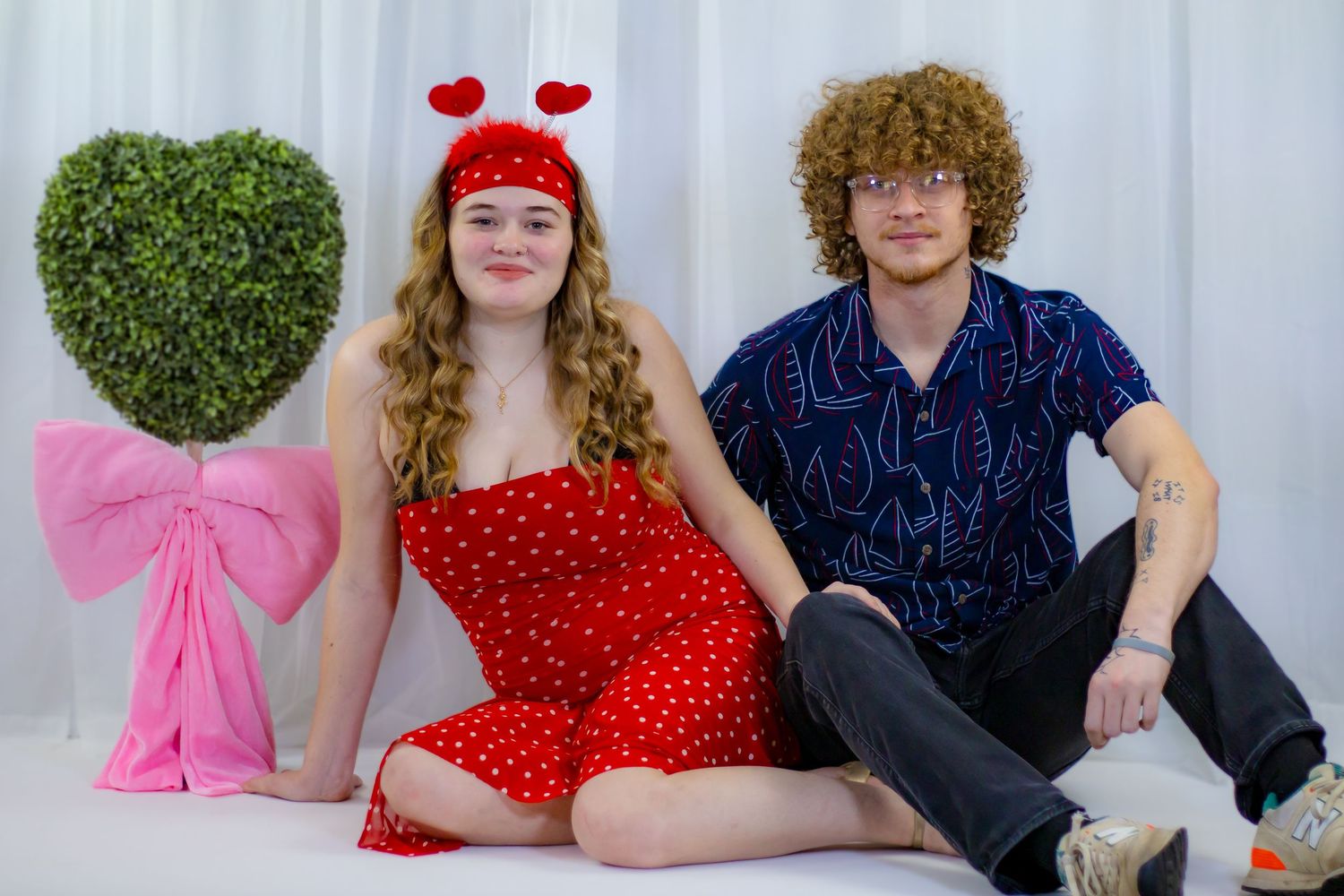 Young couple in Valentine's Day outfits sit together beside a heart-shaped topiary with pink bow.