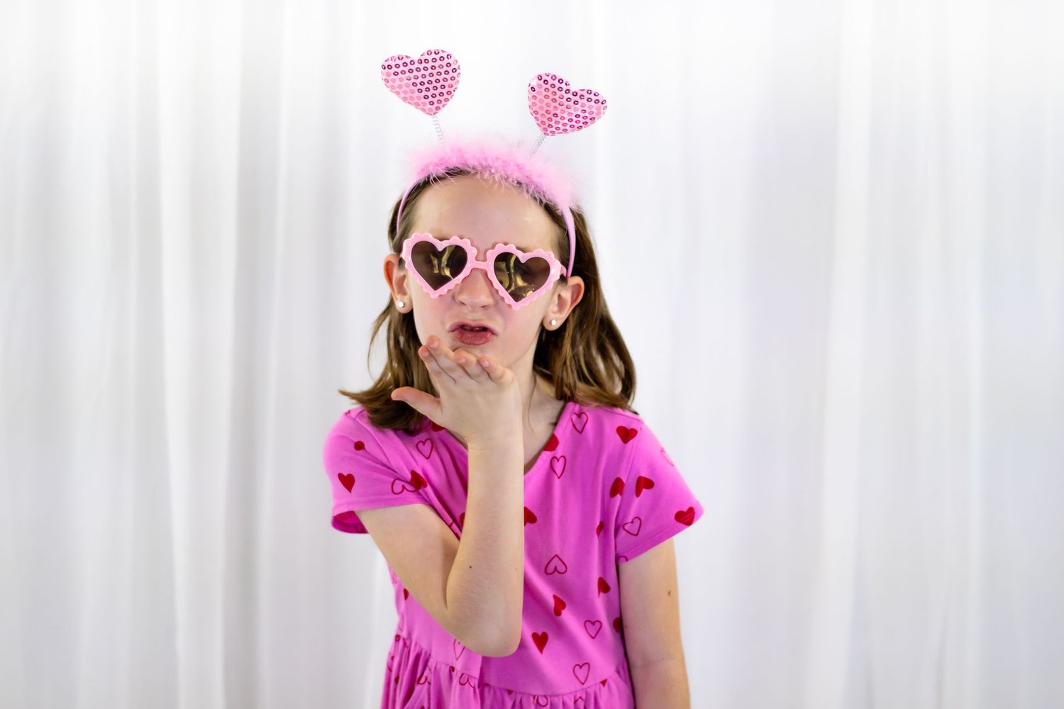Young girl in pink dress blows a kiss while wearing heart-shaped sunglasses and pink heart headband.