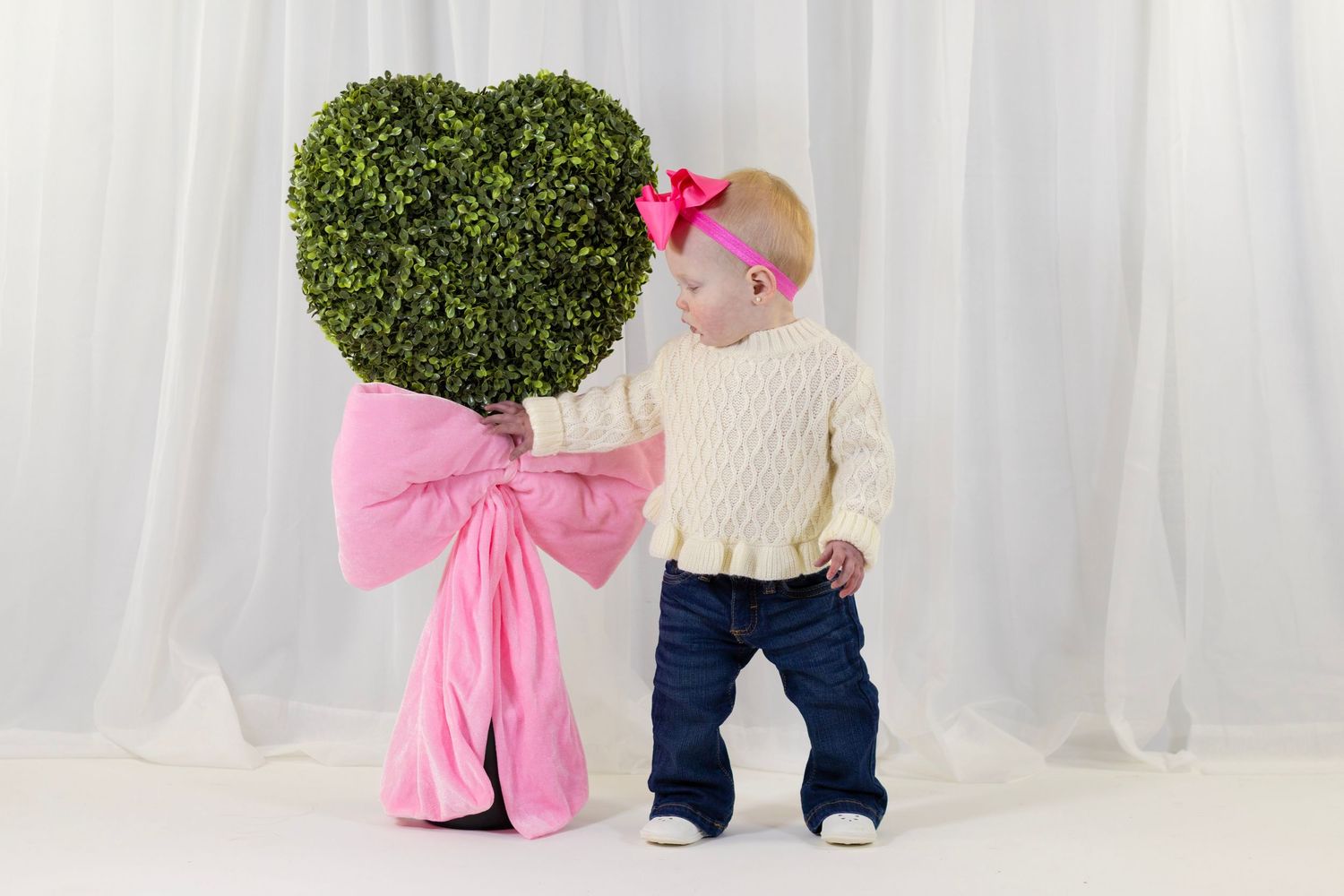 Baby girl in white sweater with pink bow standing beside a heart-shaped boxwood topiary with pink ribbon.