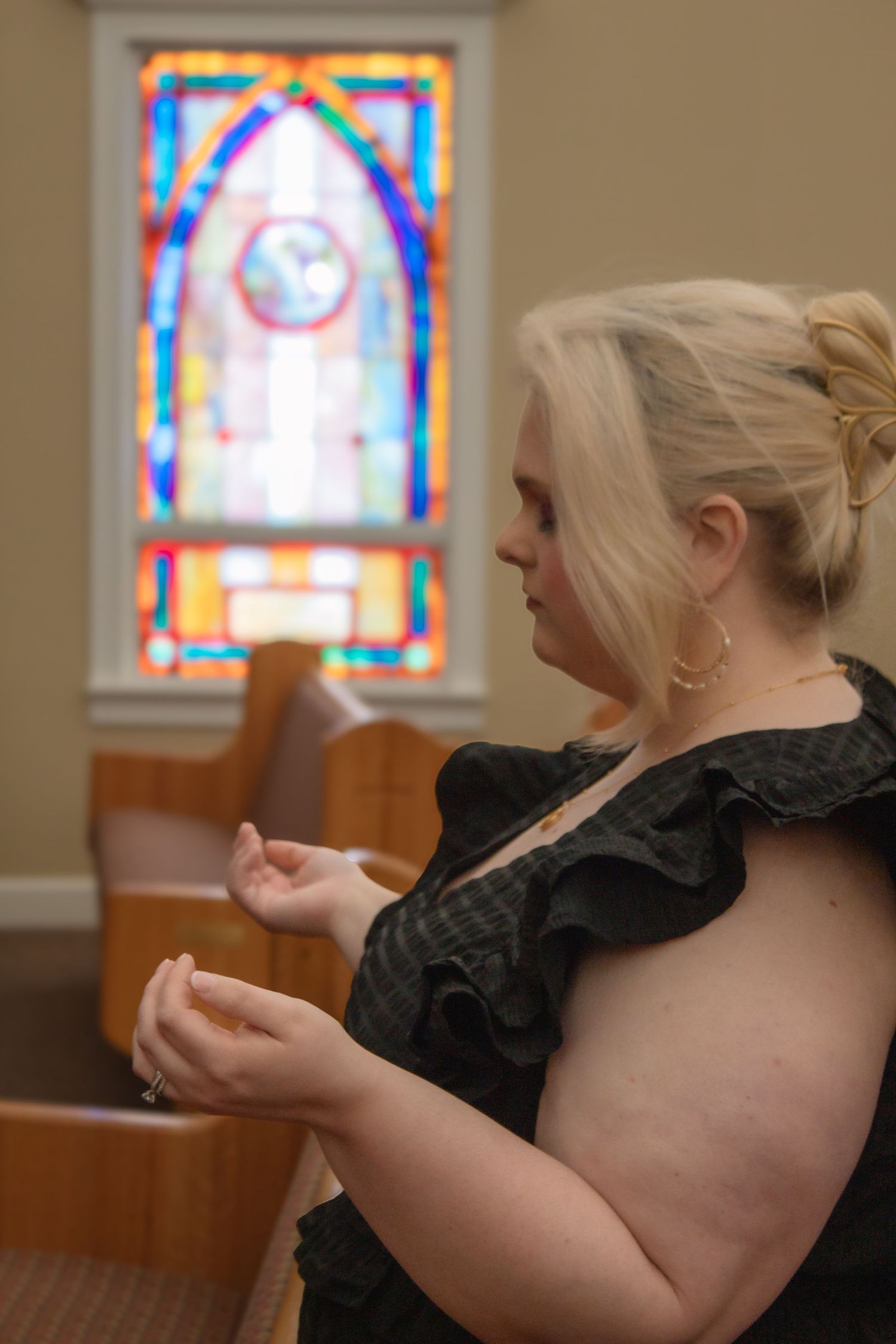 A blonde woman in a black ruffled dress prays with clasped hands near a colorful stained glass church window.