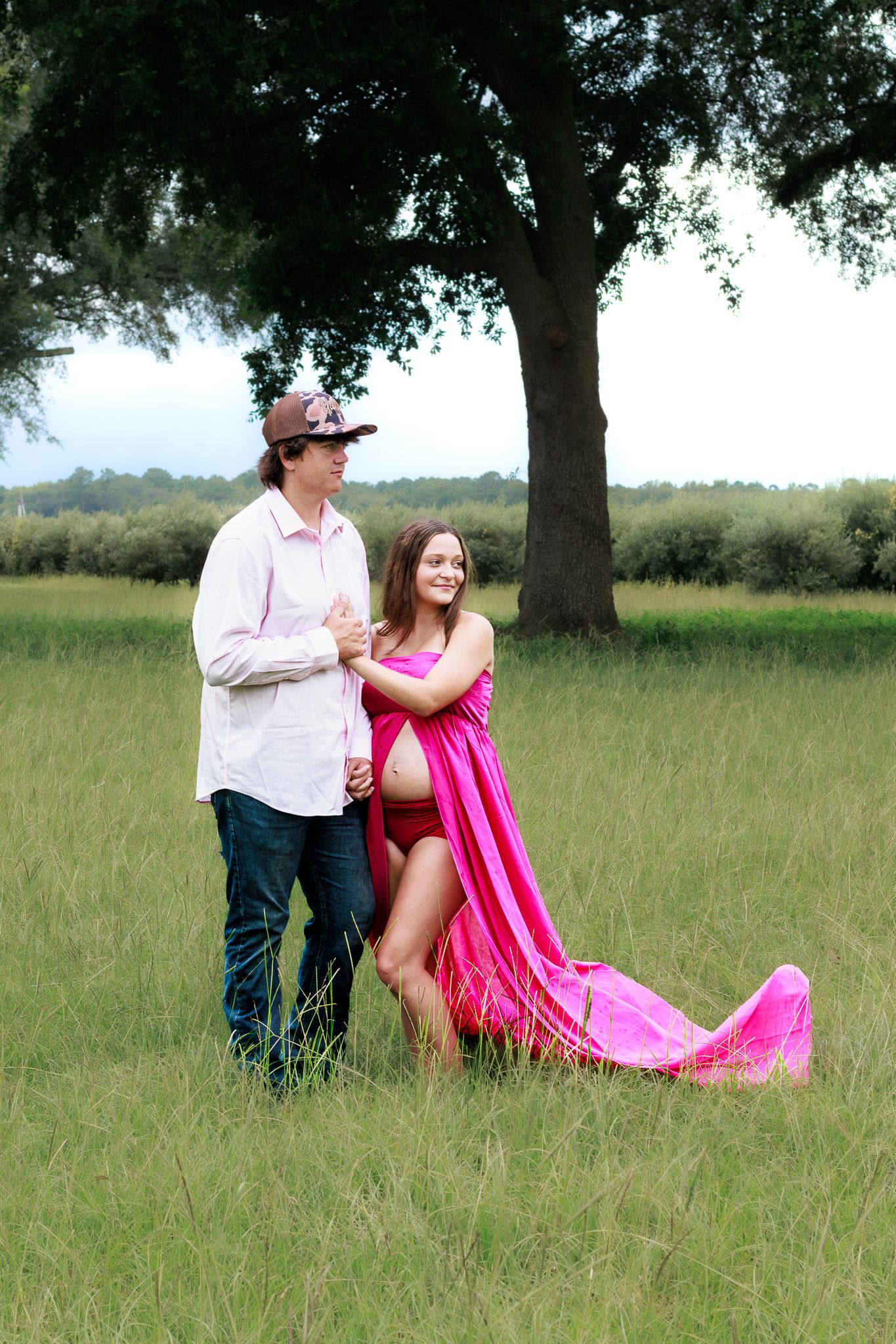 Pregnant woman in pink maternity gown poses with partner in cowboy hat in lush green field near large oak tree.