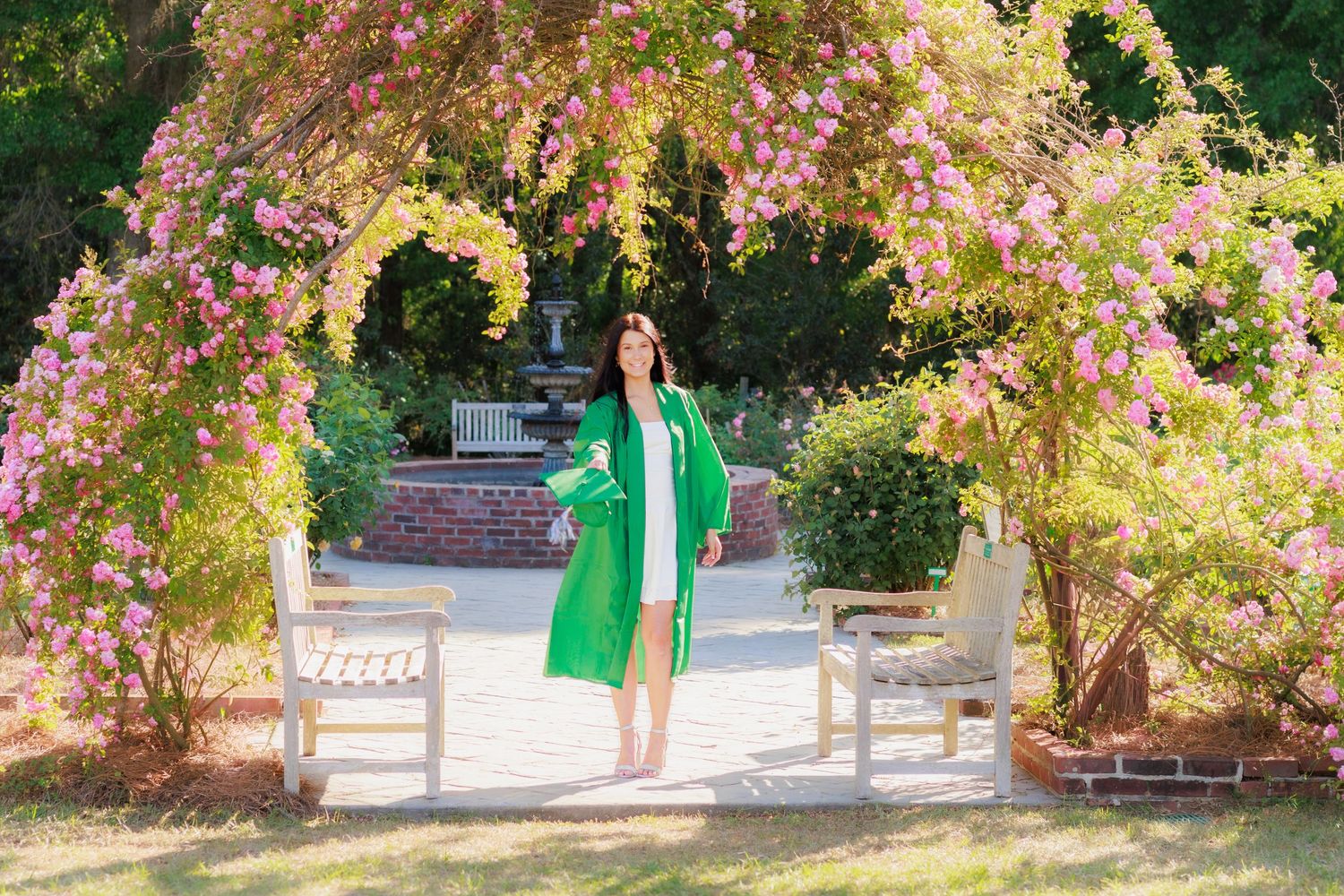 Woman in green graduation gown stands under floral arch with pink blooms in a sunny garden.