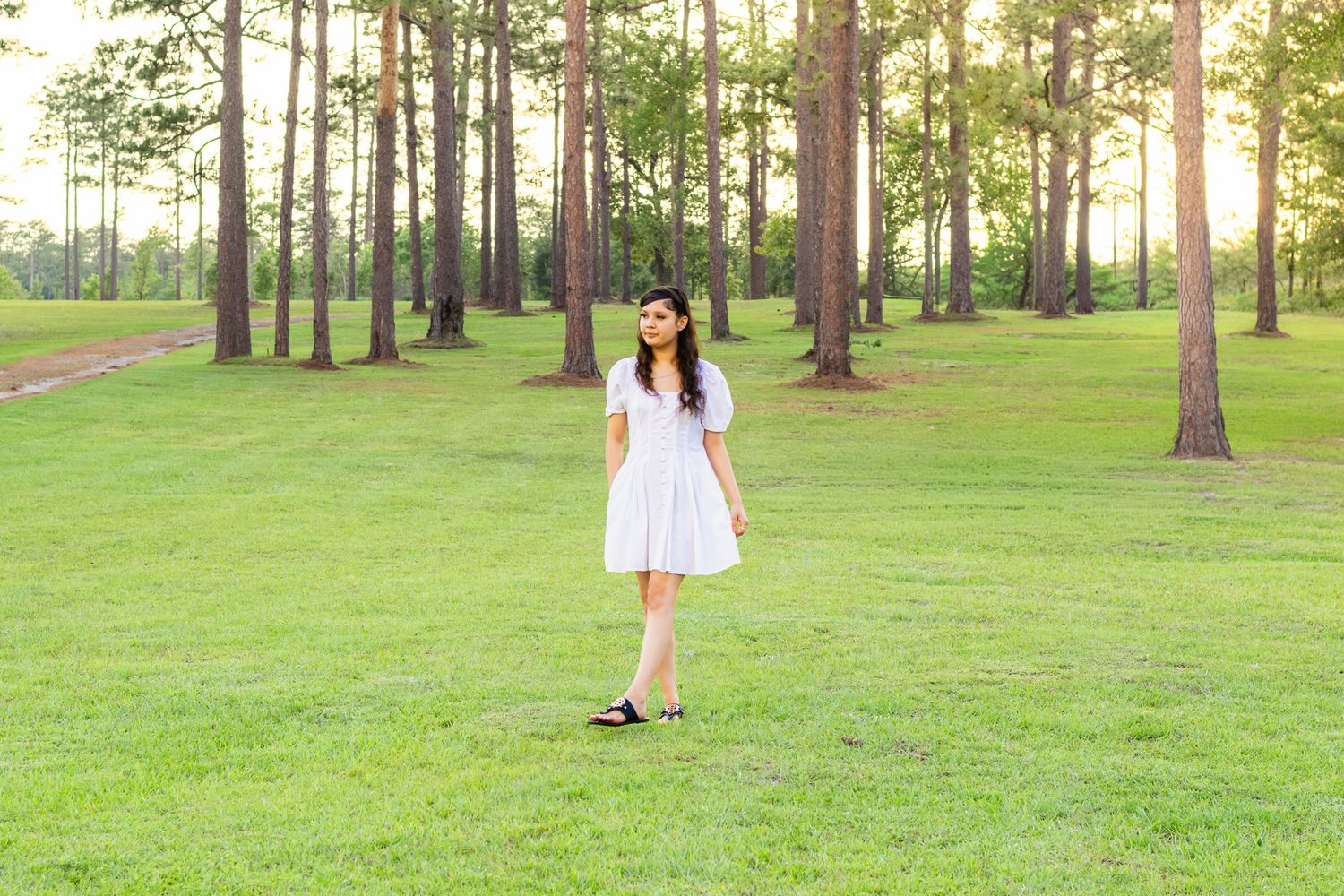 Young woman in white dress standing on lush green grass surrounded by tall pine trees in a park.