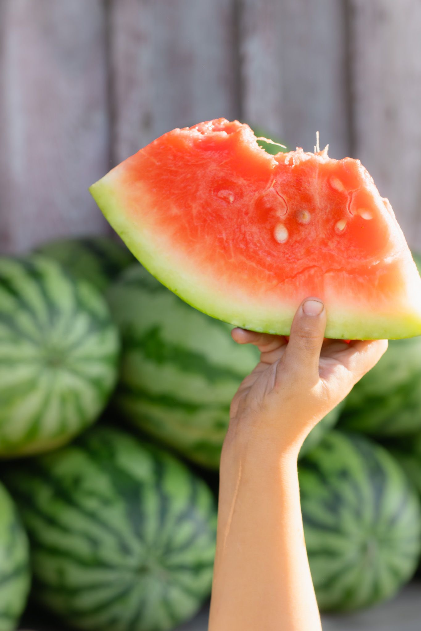 Hand holding a partially eaten slice of ripe red watermelon with whole watermelons stacked in the background.