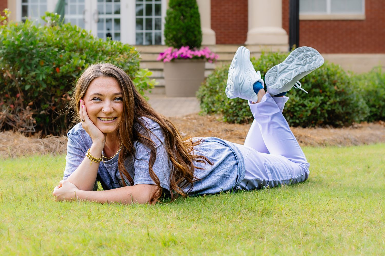 A smiling young woman with long brown hair lies on green grass in front of a brick building.