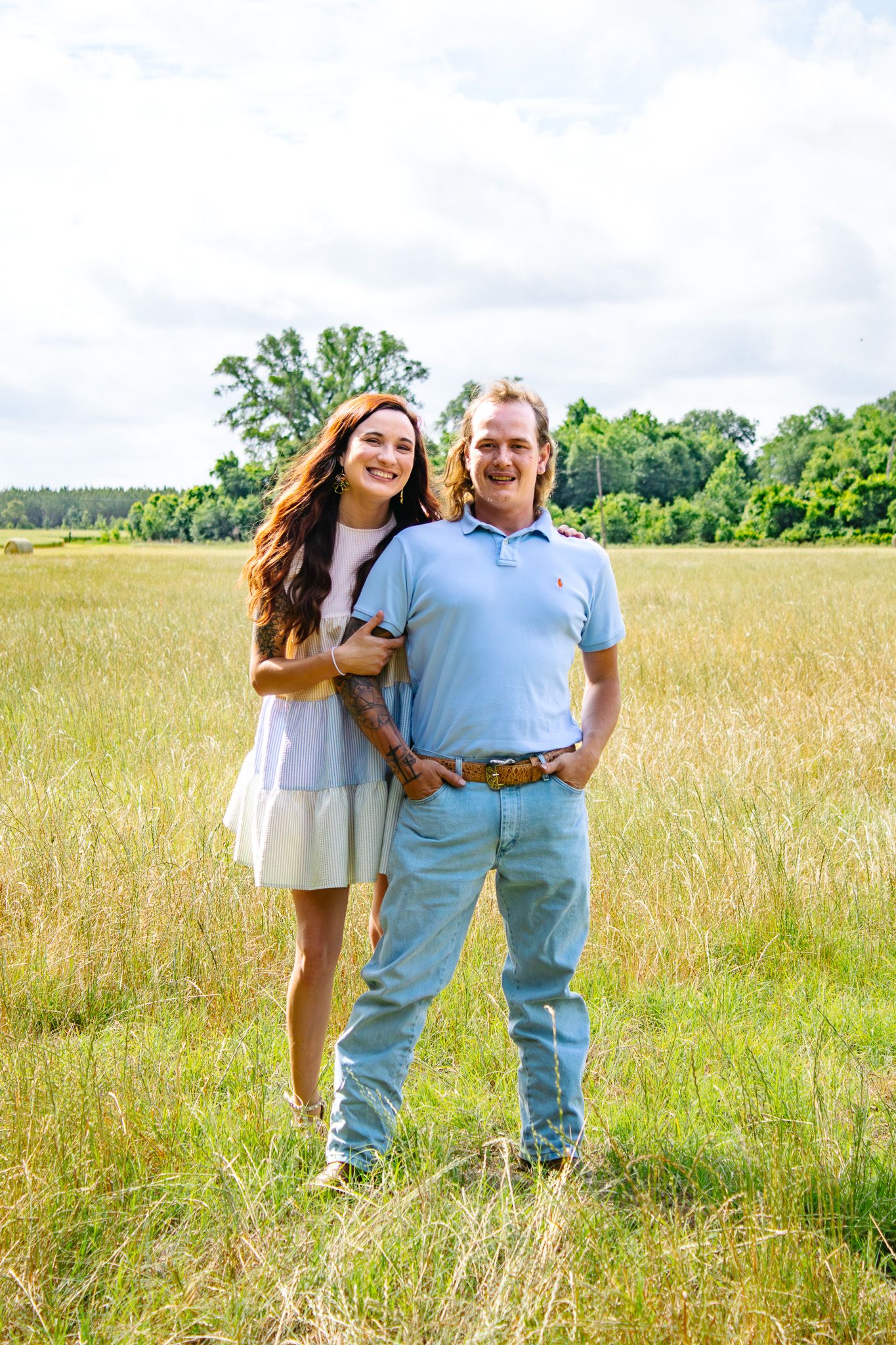 A smiling couple poses together in a golden grass field with trees and cloudy sky backdrop.