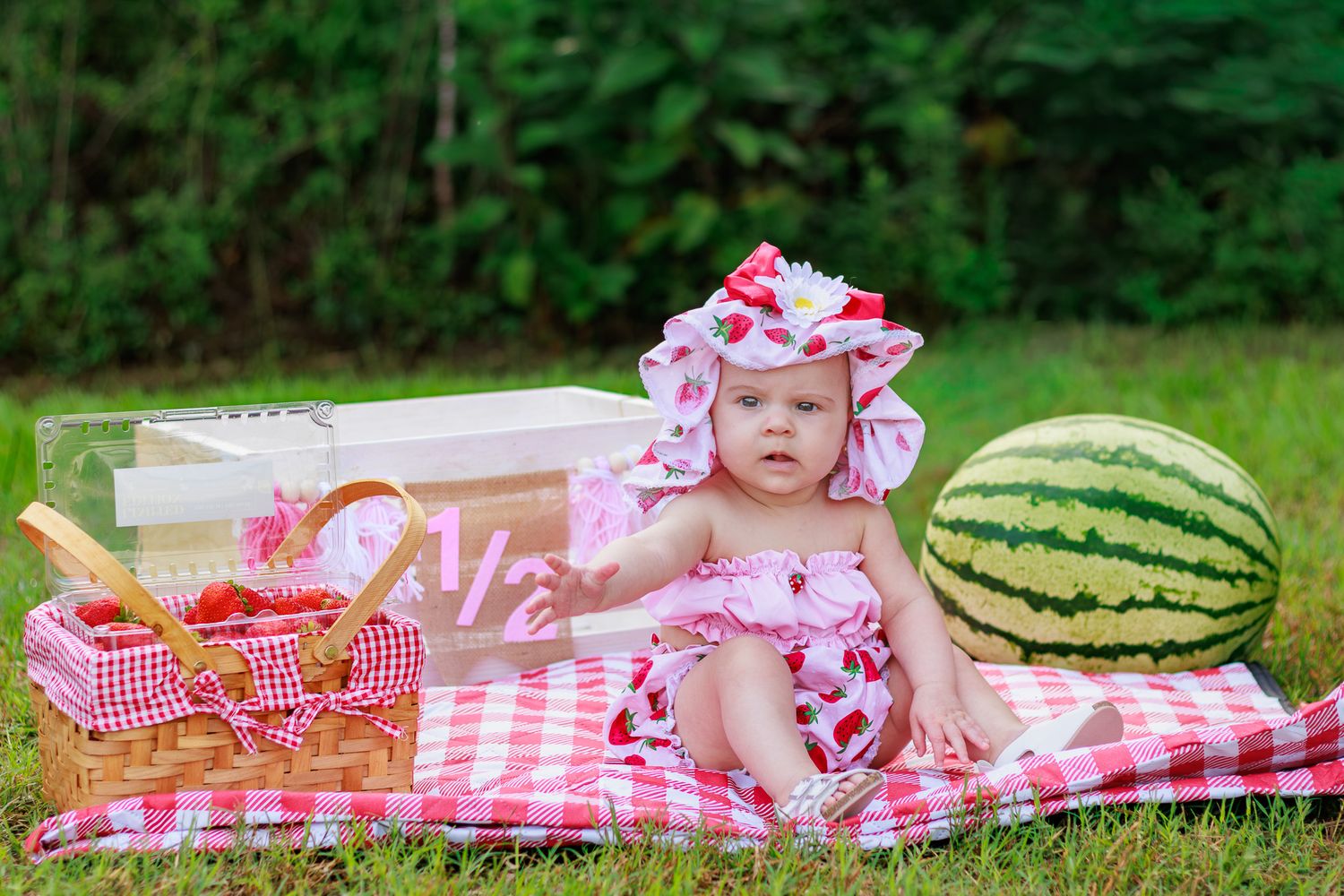Baby girl in strawberry outfit and hat sits on checkered blanket with watermelon and picnic basket outdoors.