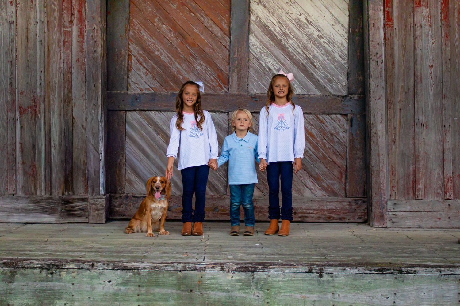 Three children and a Spaniel dog standing in front of a weathered barn door.