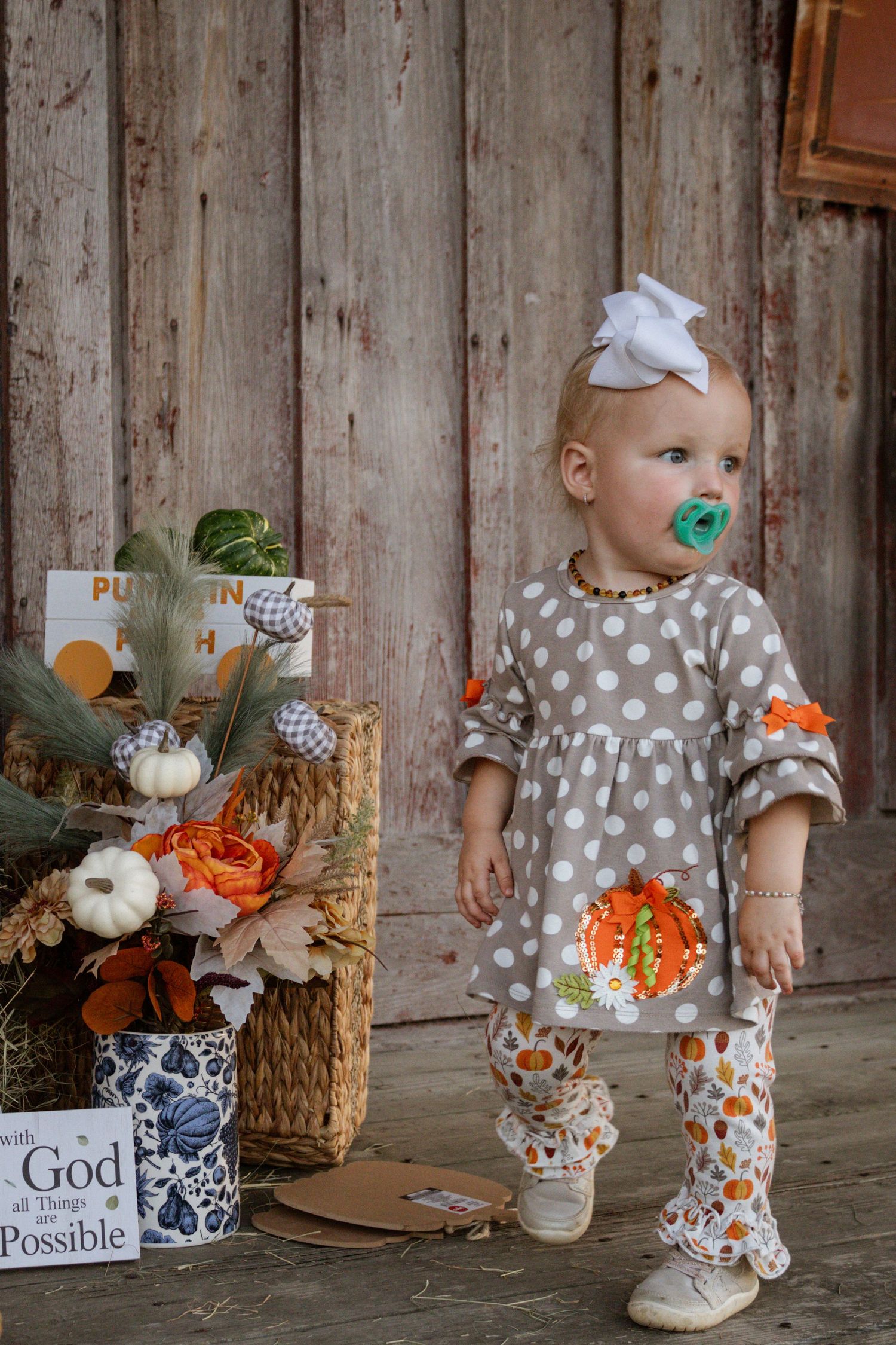 Toddler girl in polka dot pumpkin dress with pacifier standing beside fall floral arrangement.