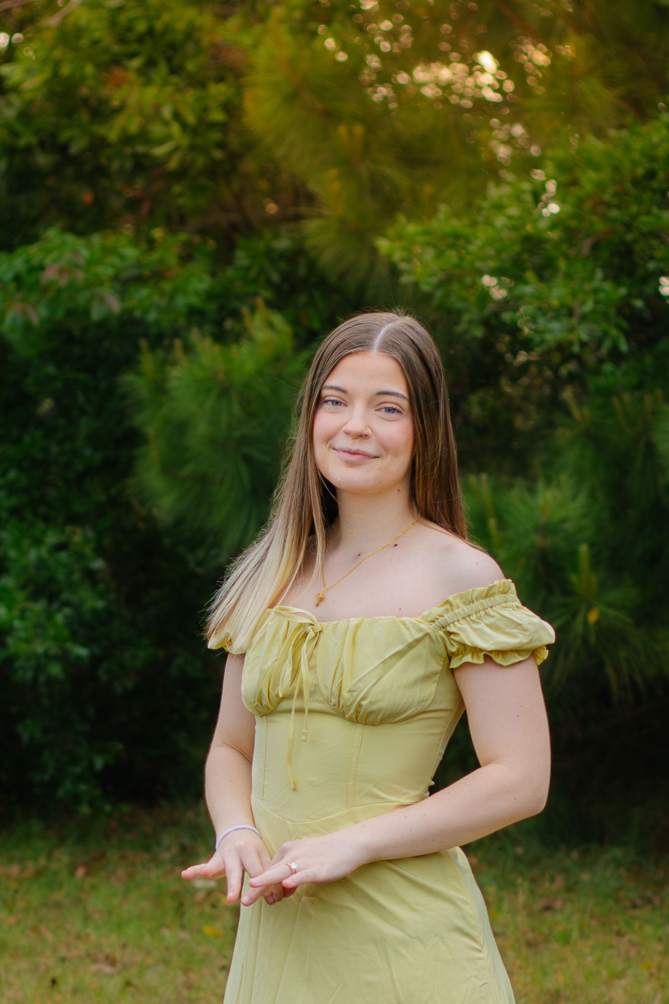 Young woman in yellow off-shoulder dress smiling outdoors with lush green trees behind her.