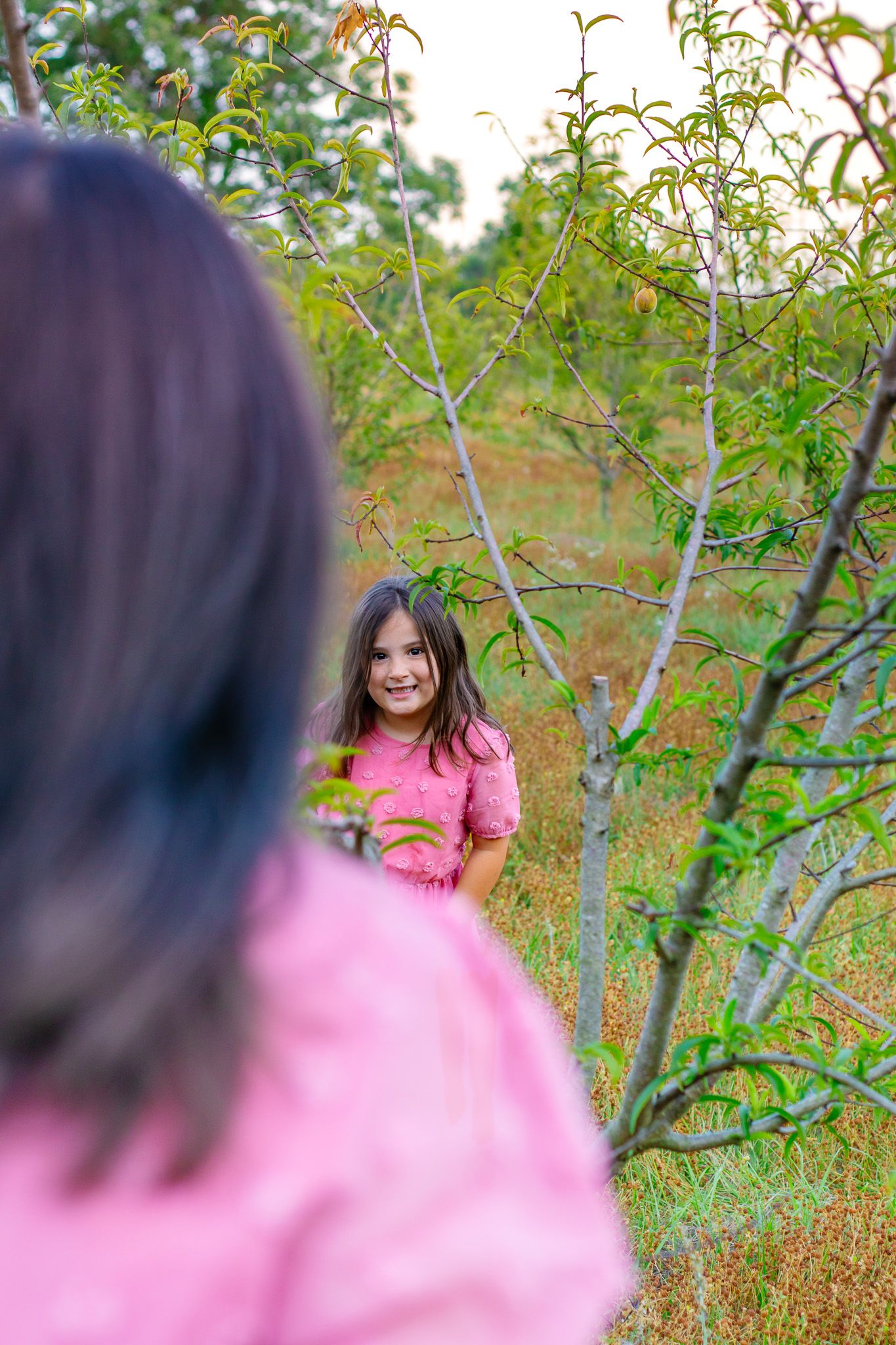 A smiling girl in a pink dress stands among trees in a field while her mother watches.