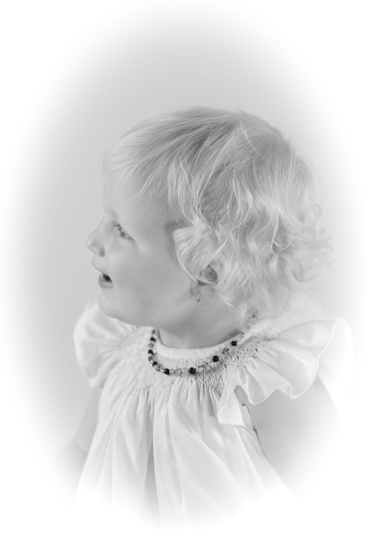 Black and white portrait of a young blonde toddler girl wearing a white smocked dress and beaded necklace.