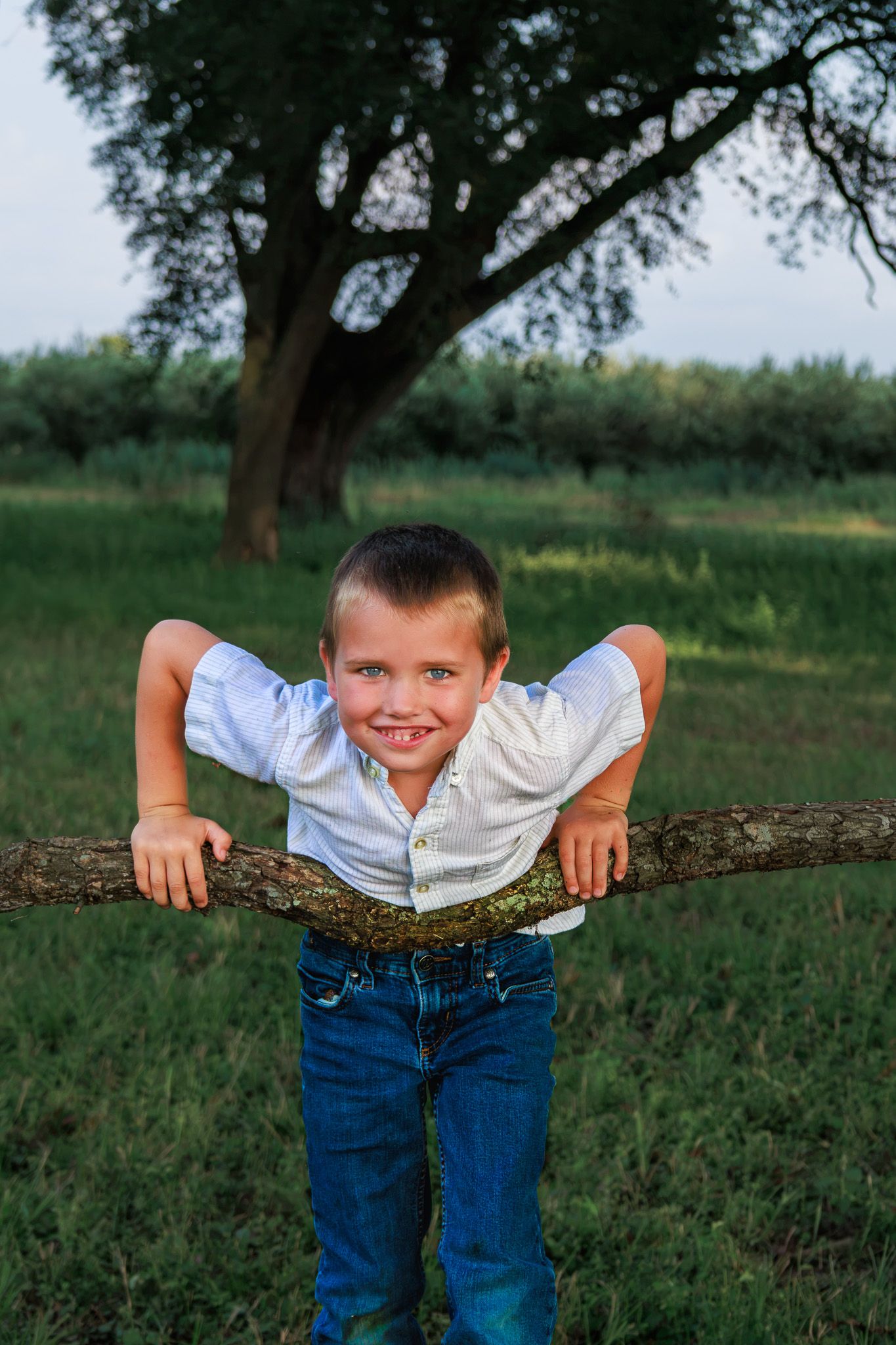 A smiling young boy in a white shirt and jeans leans over a tree branch in a lush green outdoor setting.