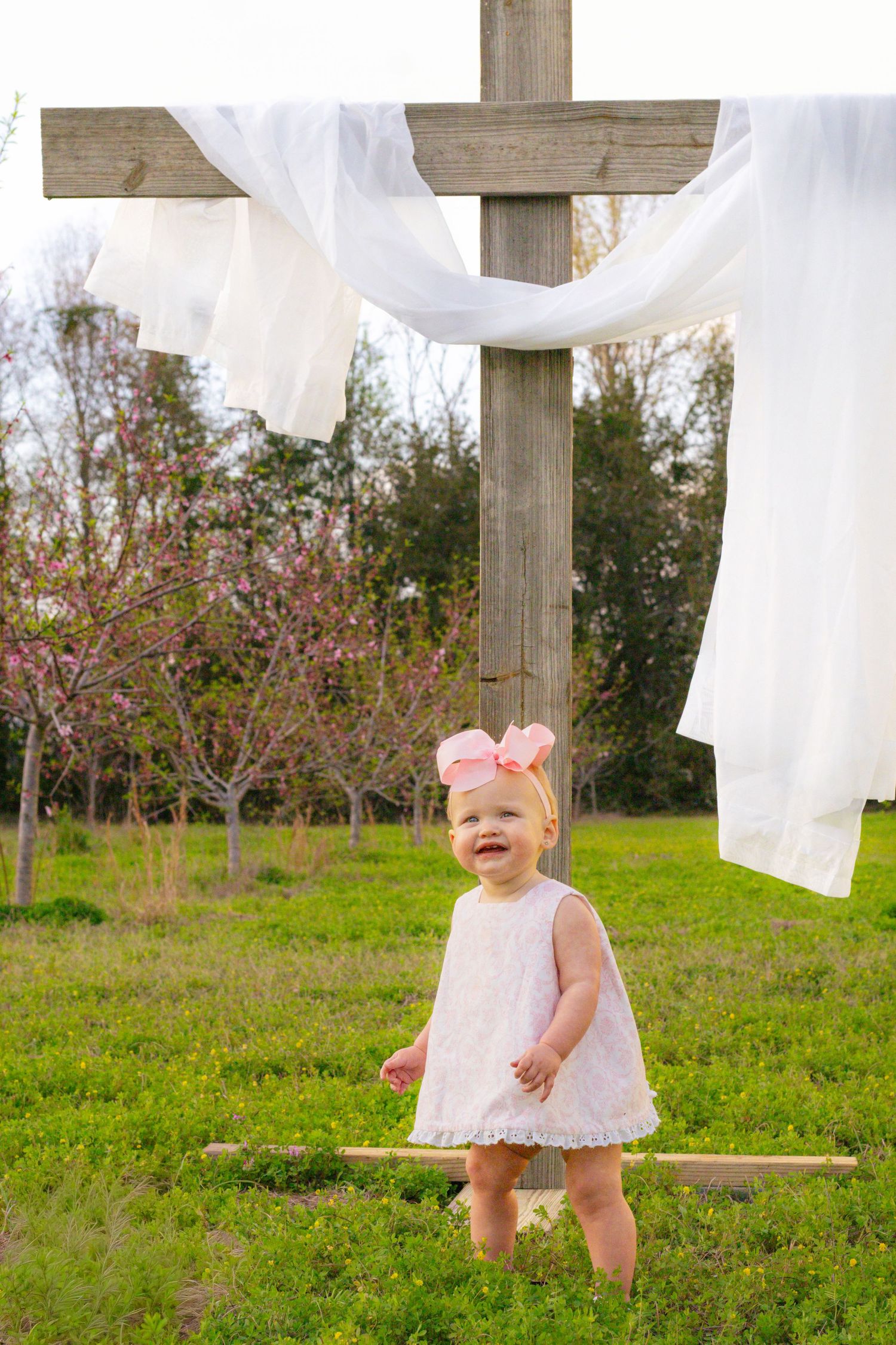 Happy baby girl in white lace dress stands laughing outdoors near a large wooden cross with white draping.