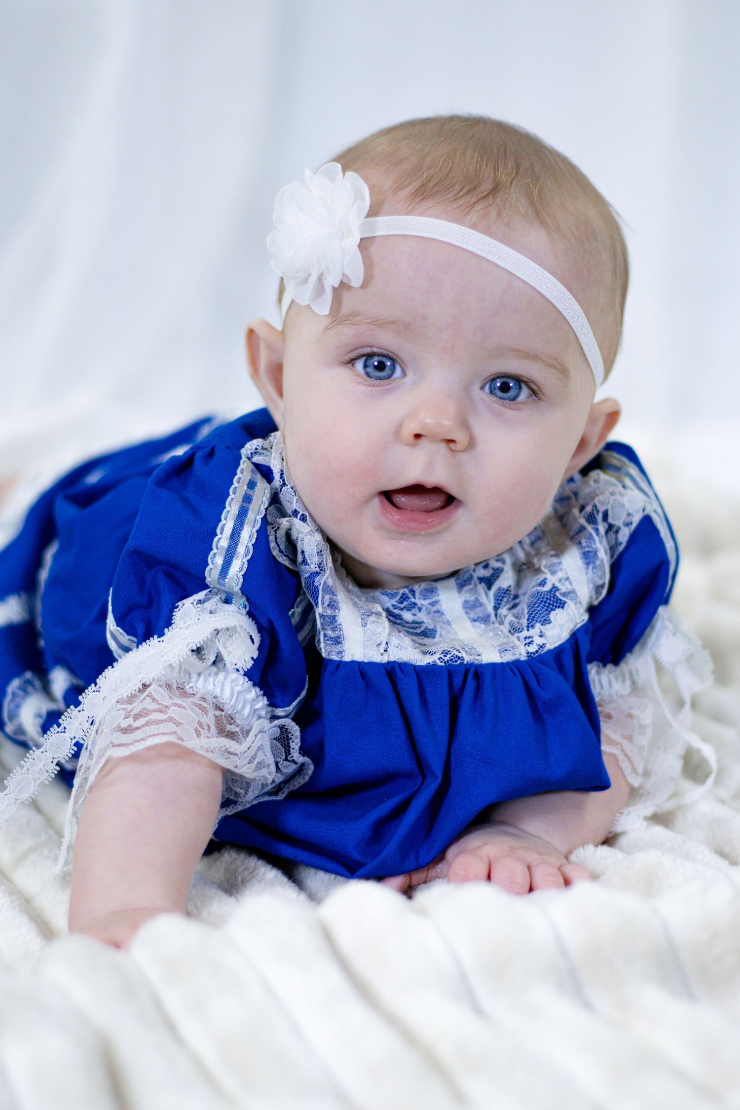 Adorable baby girl in blue lace dress with white flower headband on cream blanket.