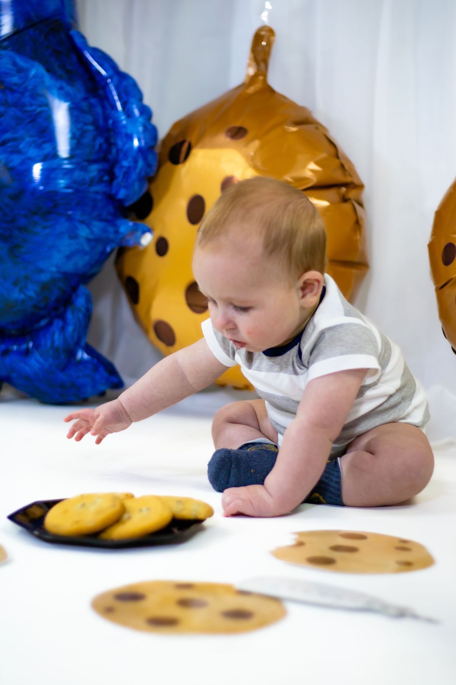 Curious baby boy reaching for cookies on plate surrounded by Cookie Monster Sesame Street balloons.