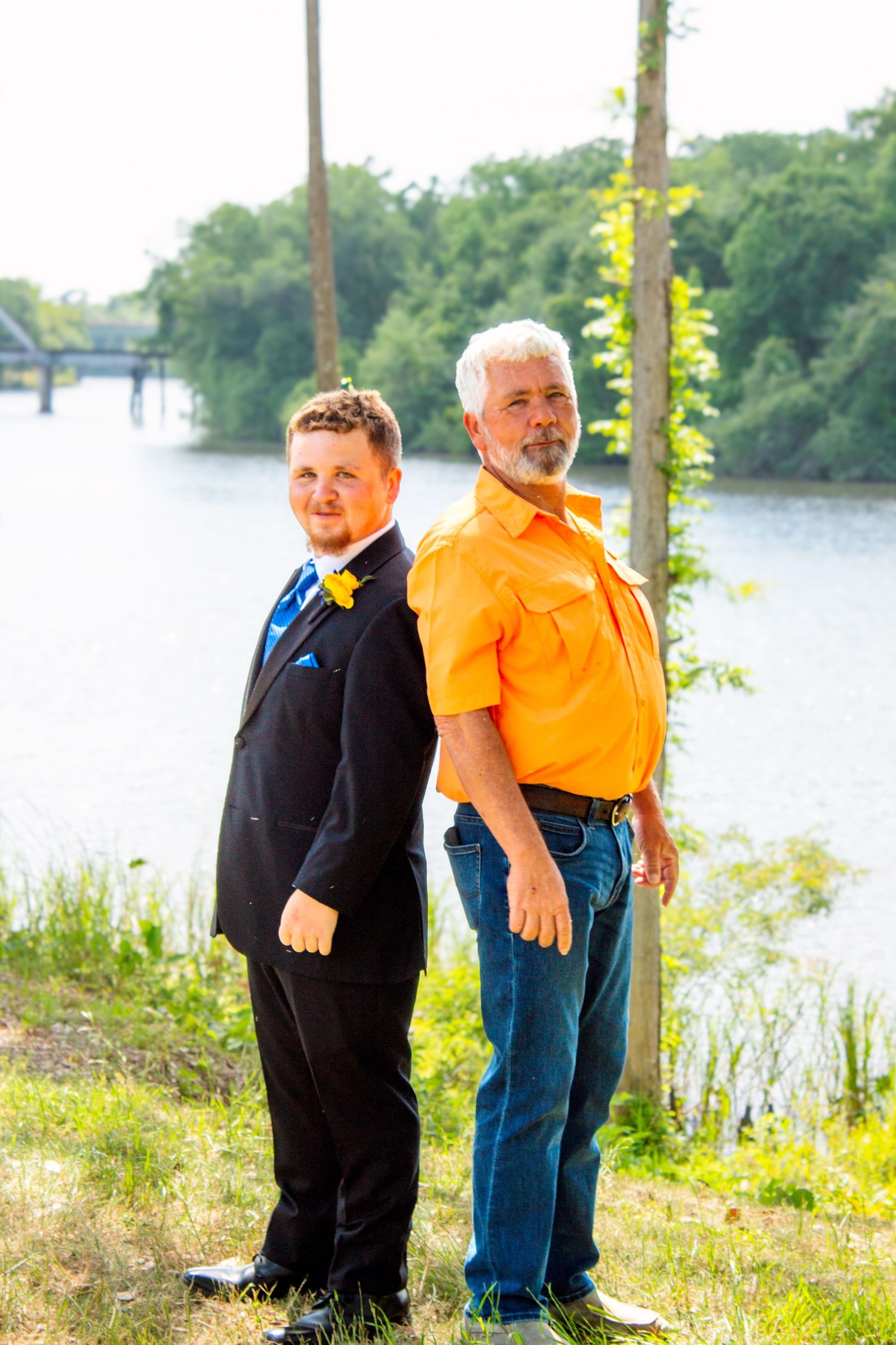 Young man in black suit with blue boutonniere stands back-to-back with older man in orange shirt outdoors.